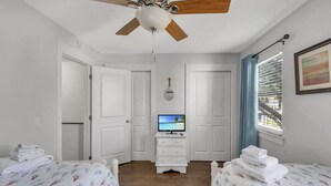 Une chambre d’hôtel avec deux lits, un ventilateur de plafond, un téléviseur posé sur un meuble et une fenêtre avec des rideaux.