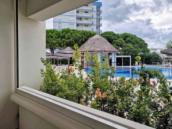 Zona de piscina con una estructura de techo de paja, rodeada de vegetación y un edificio de varios pisos al fondo.