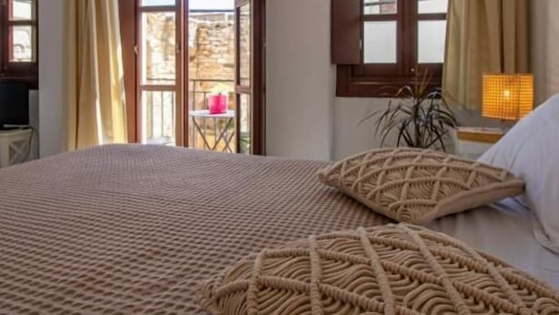 A bedroom with a large bed, two windows with curtains, a small table with a lamp, and a view of the outside. - Platsa's House (Naxos)