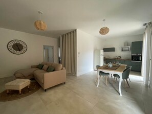 A modern living room with a beige sofa, a dining table with chairs, a kitchen area with a microwave and oven, and a large wall clock.