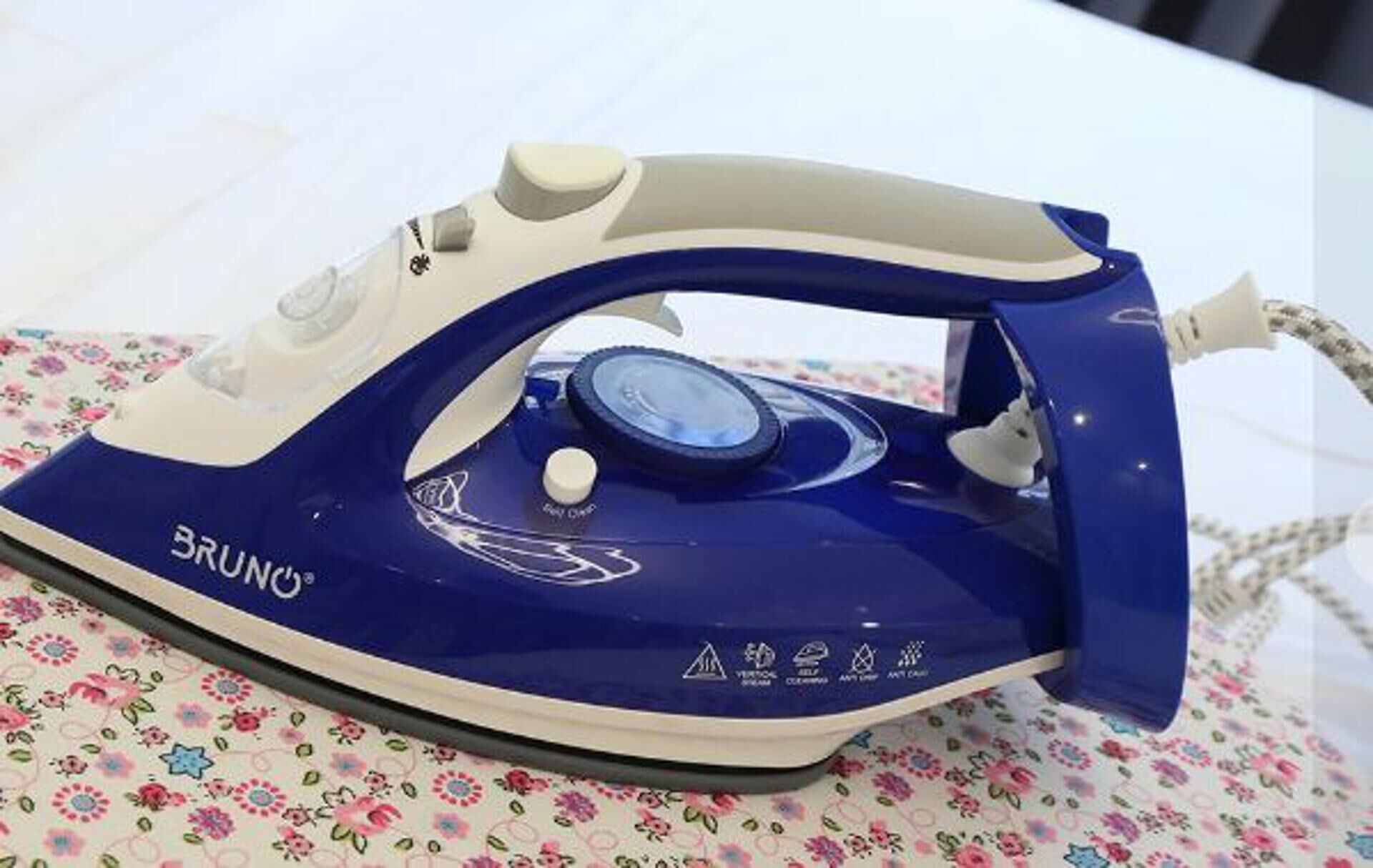 Un fer à repasser bleu et blanc, portant la marque « BRUNO », est posé sur une nappe à motifs floraux.