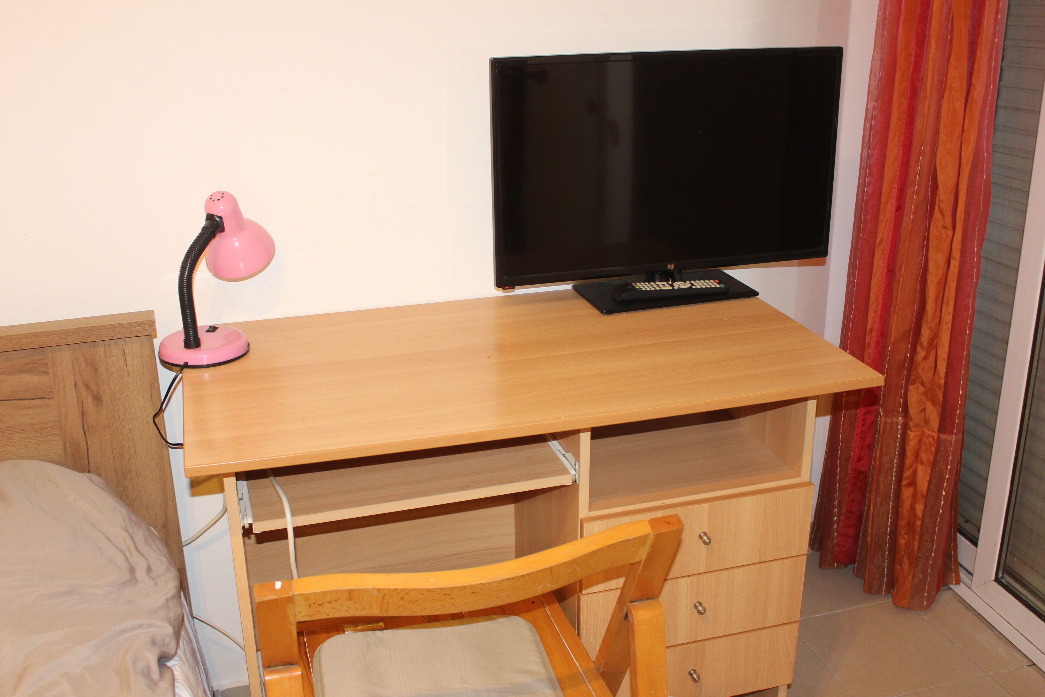 Un dormitorio con un escritorio de madera, una lámpara de escritorio rosa, un televisor, una silla de madera y una cama.