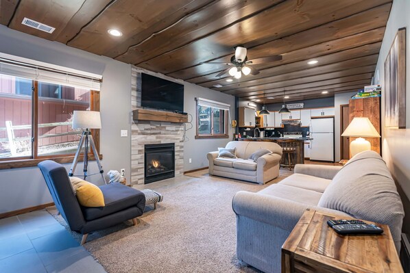 A cozy living room with a fireplace, a sofa, a recliner, and a dining area with a kitchen in the background.