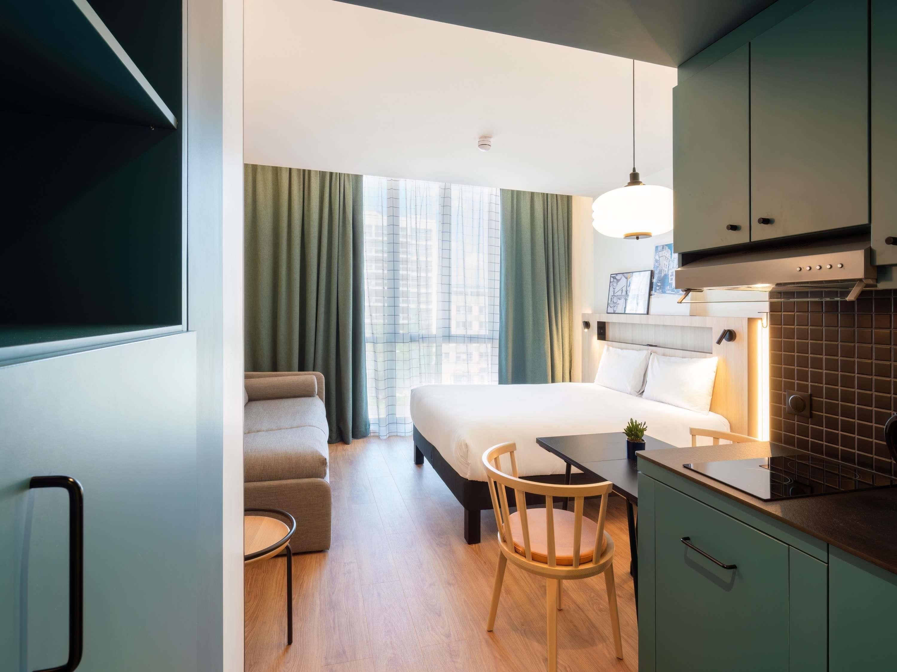 A modern hotel room with a bed, a desk, a chair, and a kitchenette.