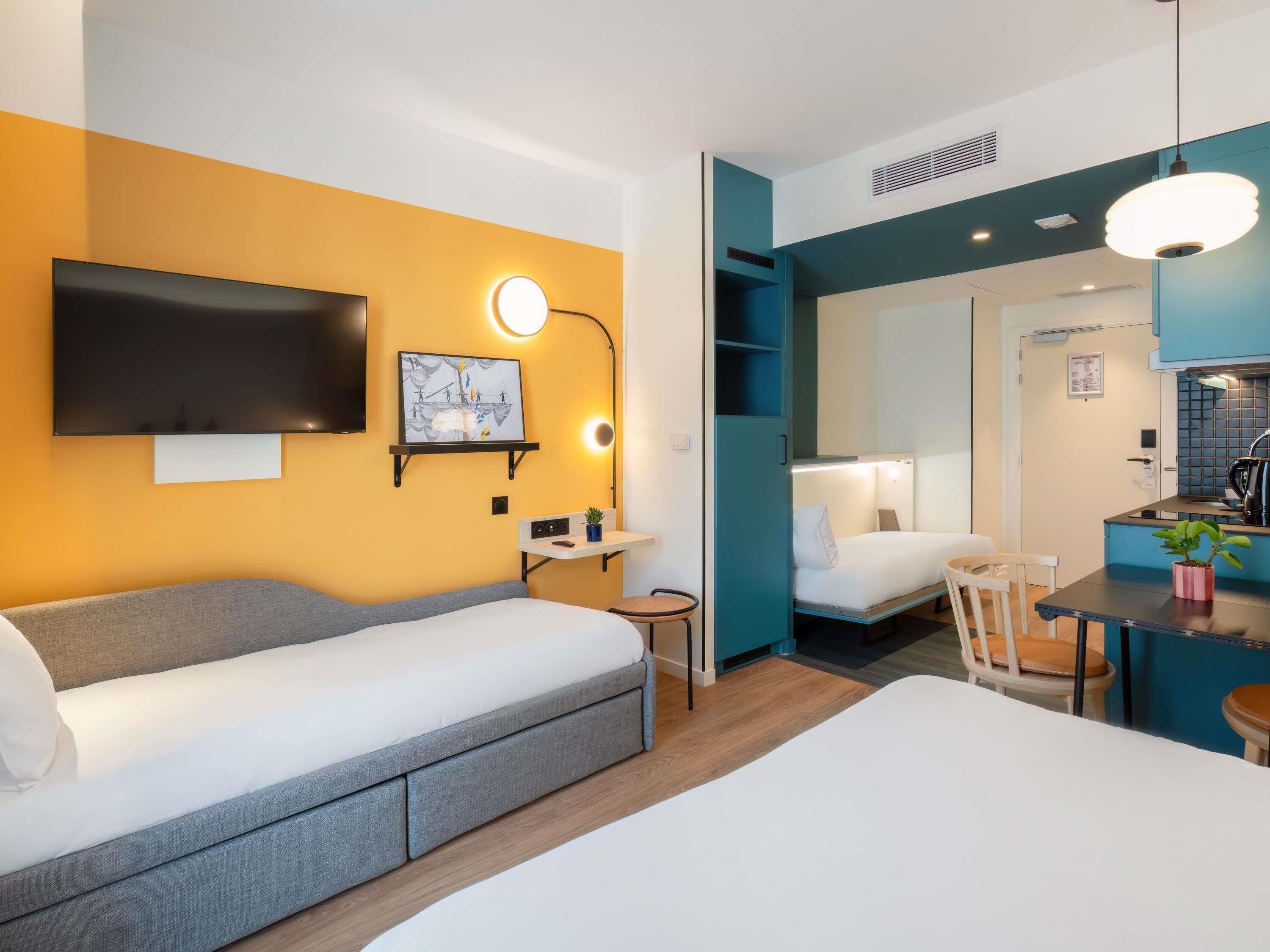 A modern hotel room with a sofa bed, a small desk with a lamp, a TV mounted on the wall, and a kitchenette area.