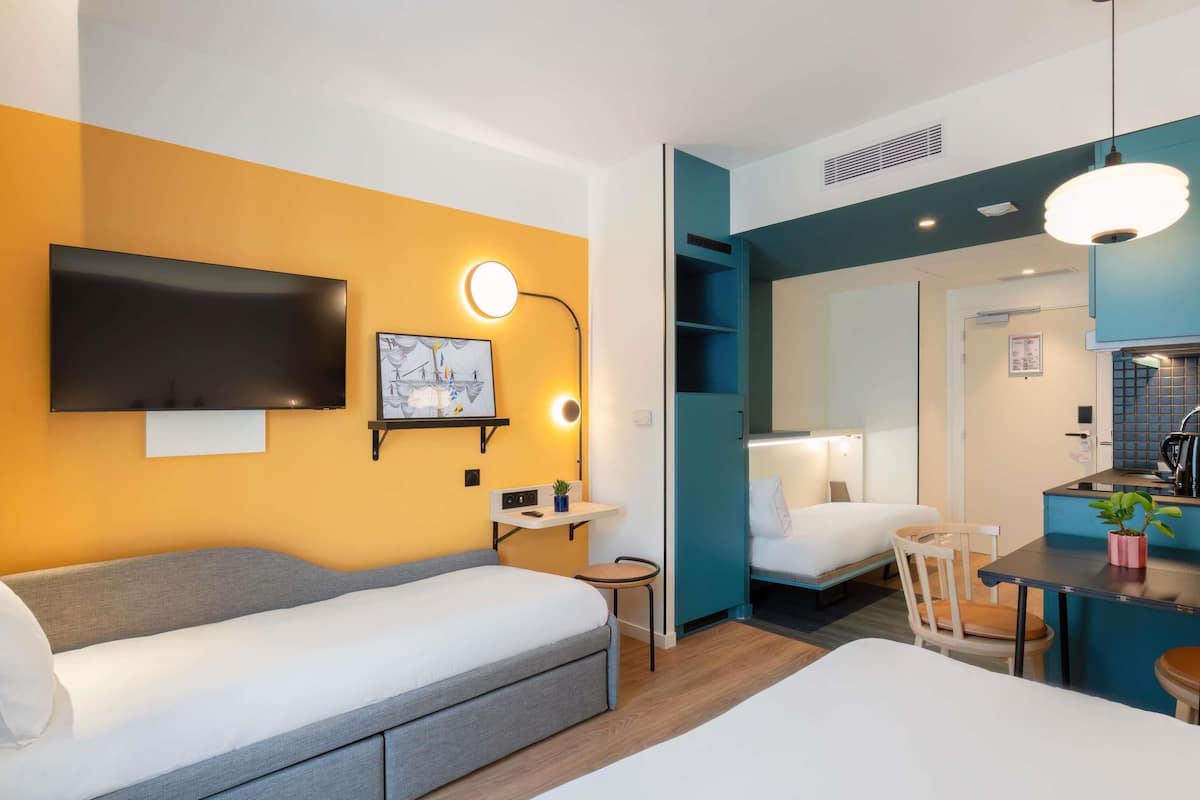 A modern hotel room with a sofa bed, a small desk with a lamp, a TV mounted on the wall, and a kitchenette area.