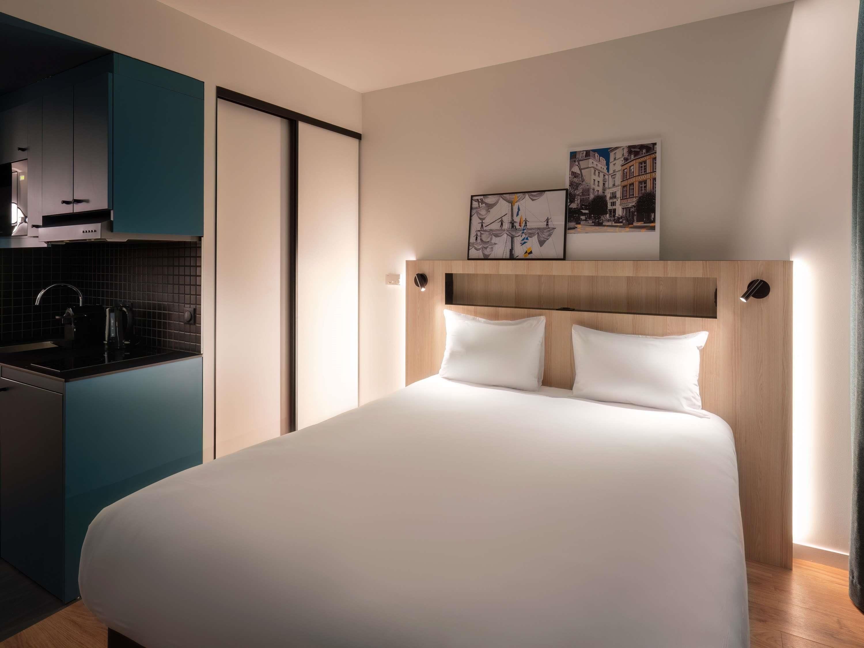 A hotel room with a bed, a kitchenette, and two framed pictures on the wall.