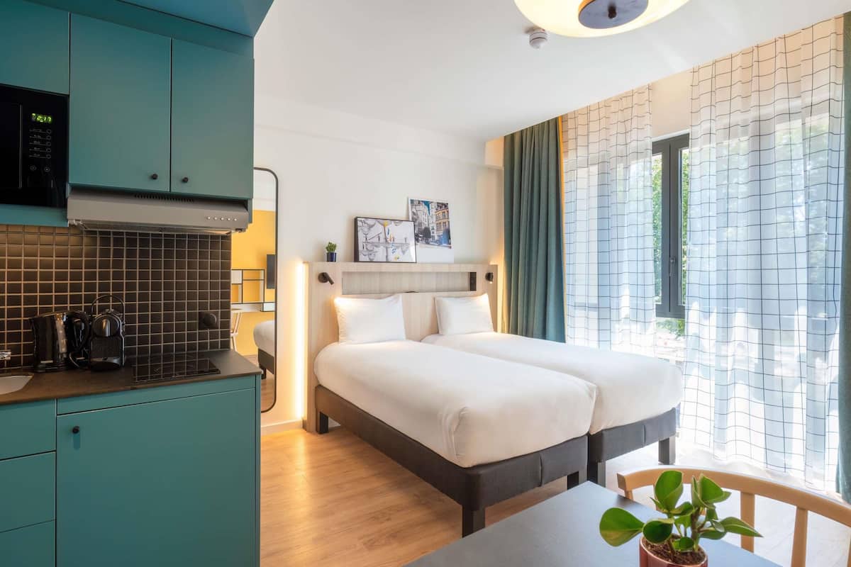 A modern hotel room with a kitchenette, two beds, a dining table, and a large window with curtains.