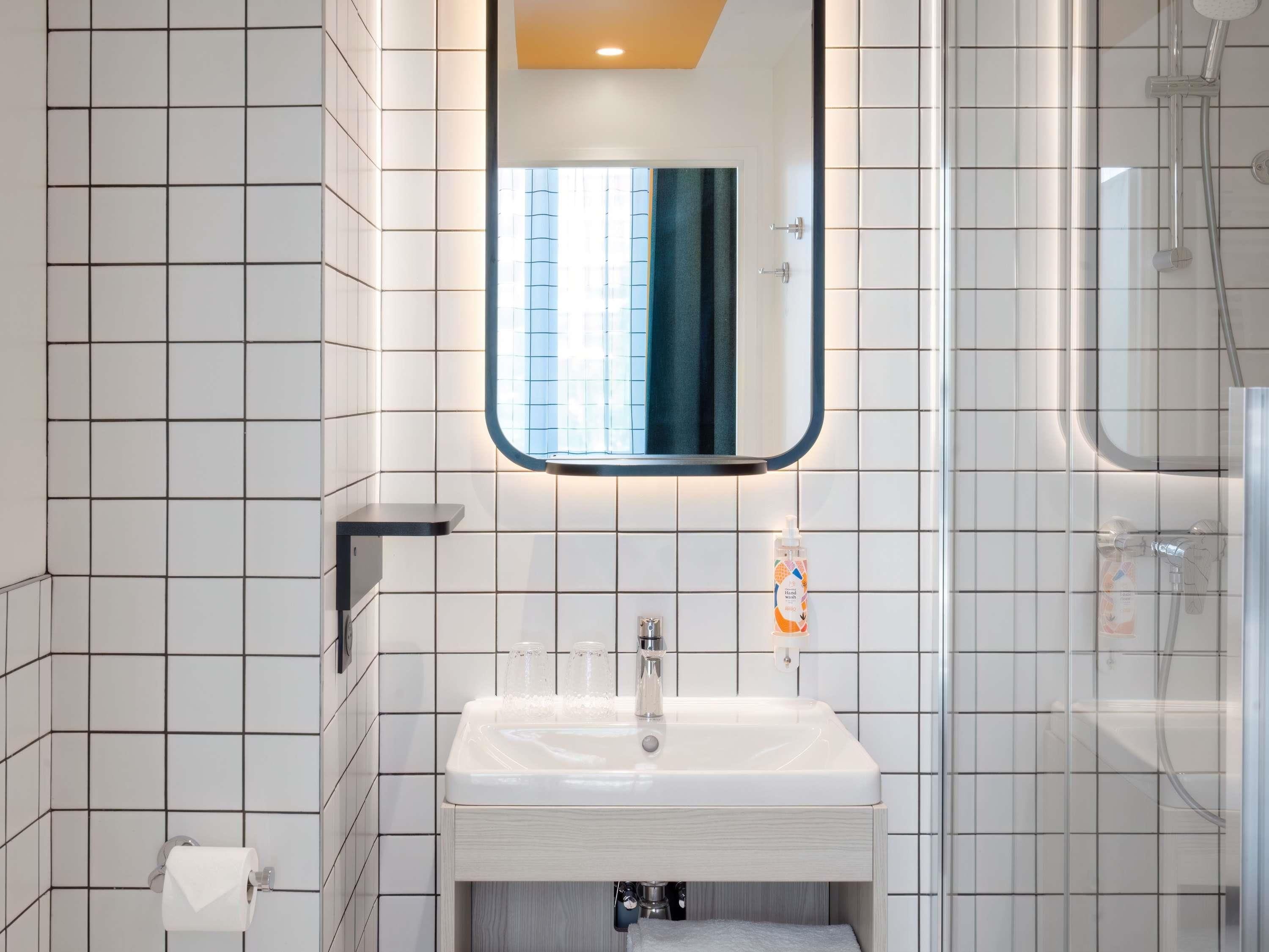 Un baño con paredes de azulejos blancos, un lavamanos blanco, un espejo, un perchero para toallas y una zona de ducha.