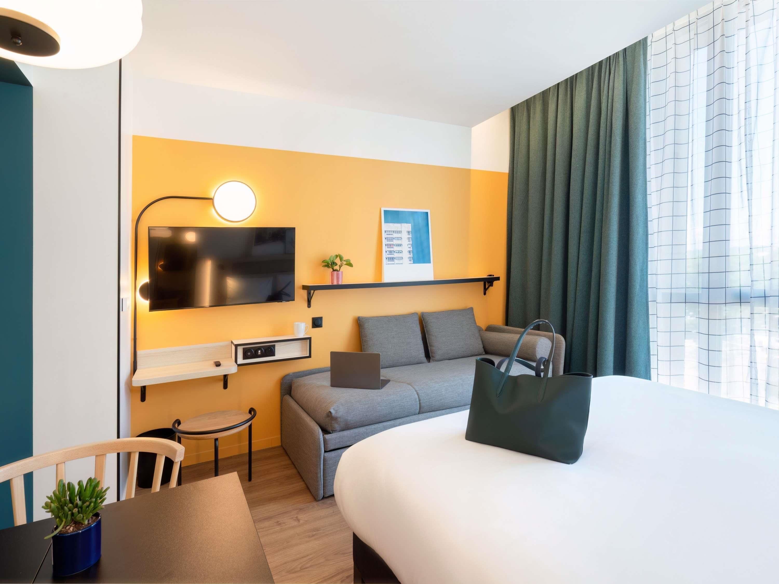 A hotel room with a bed, a desk, a chair, a TV, a shelf with a plant, and a window with curtains.