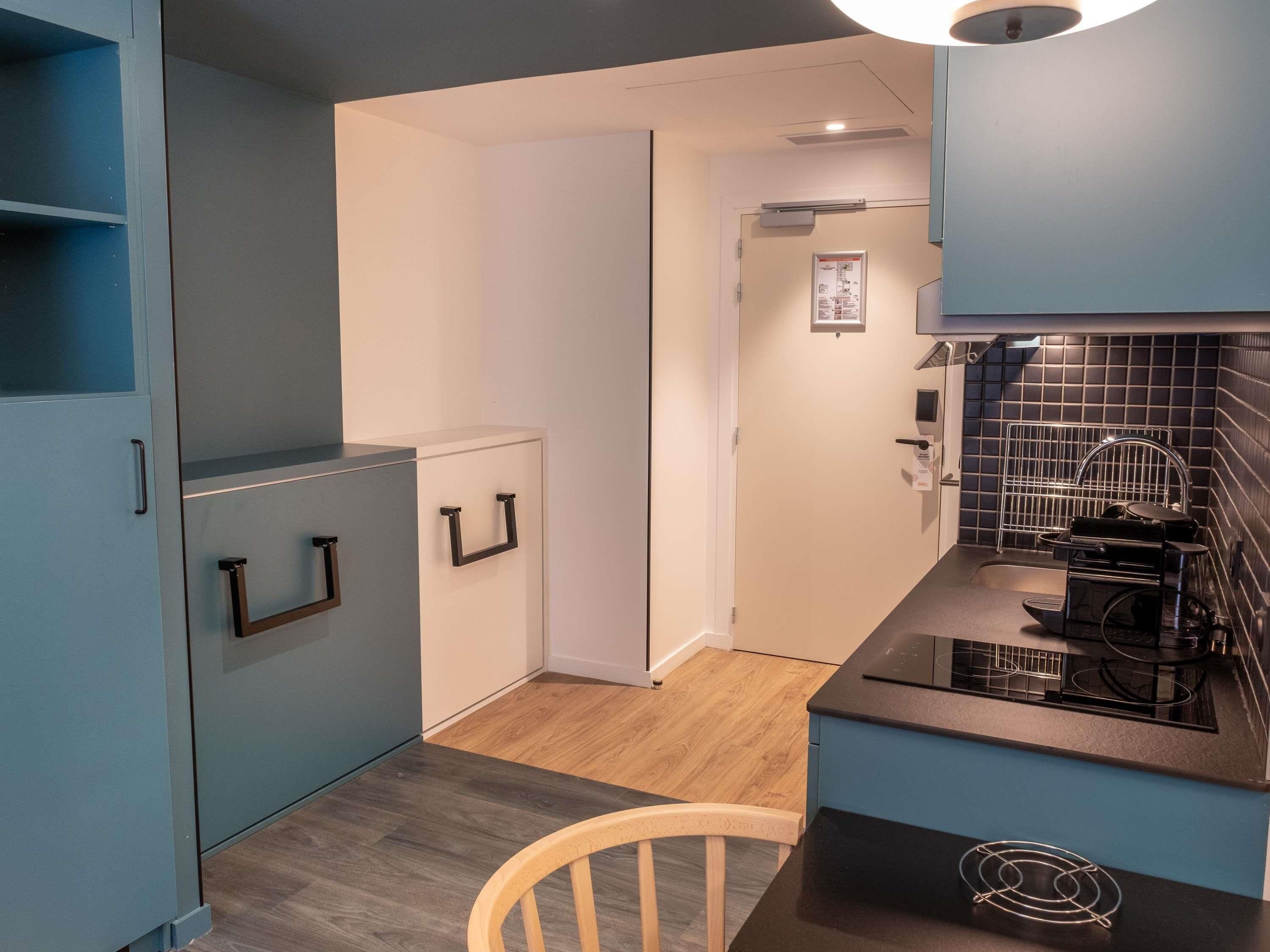 Een compacte keuken met een inbouwfornuis, een magnetron en een kleine koelkast. Er staat een houten stoel en is een deur naar een andere kamer.