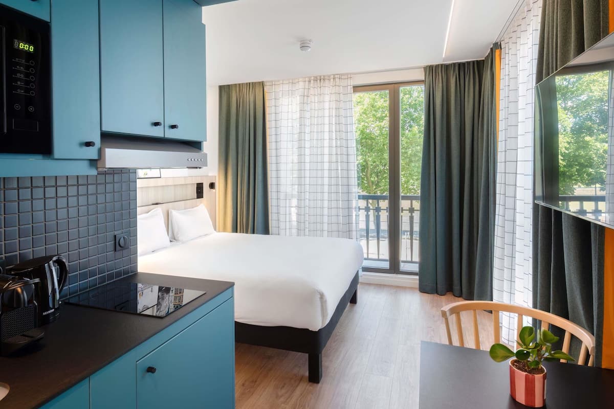 A modern hotel room with a bed, a kitchenette, a dining area, and a balcony with curtains.