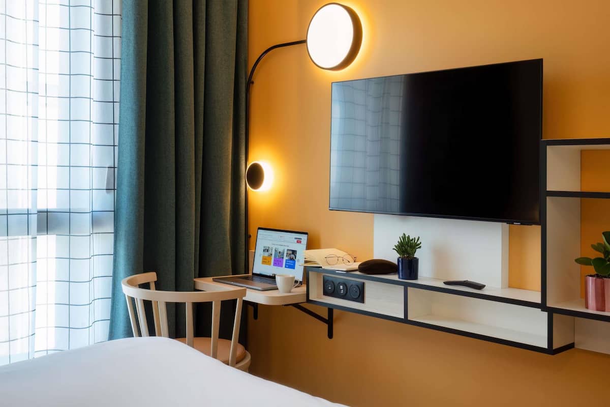 A hotel room with a bed, a desk with a laptop, a flat-screen TV, a wall-mounted light, and a potted plant.