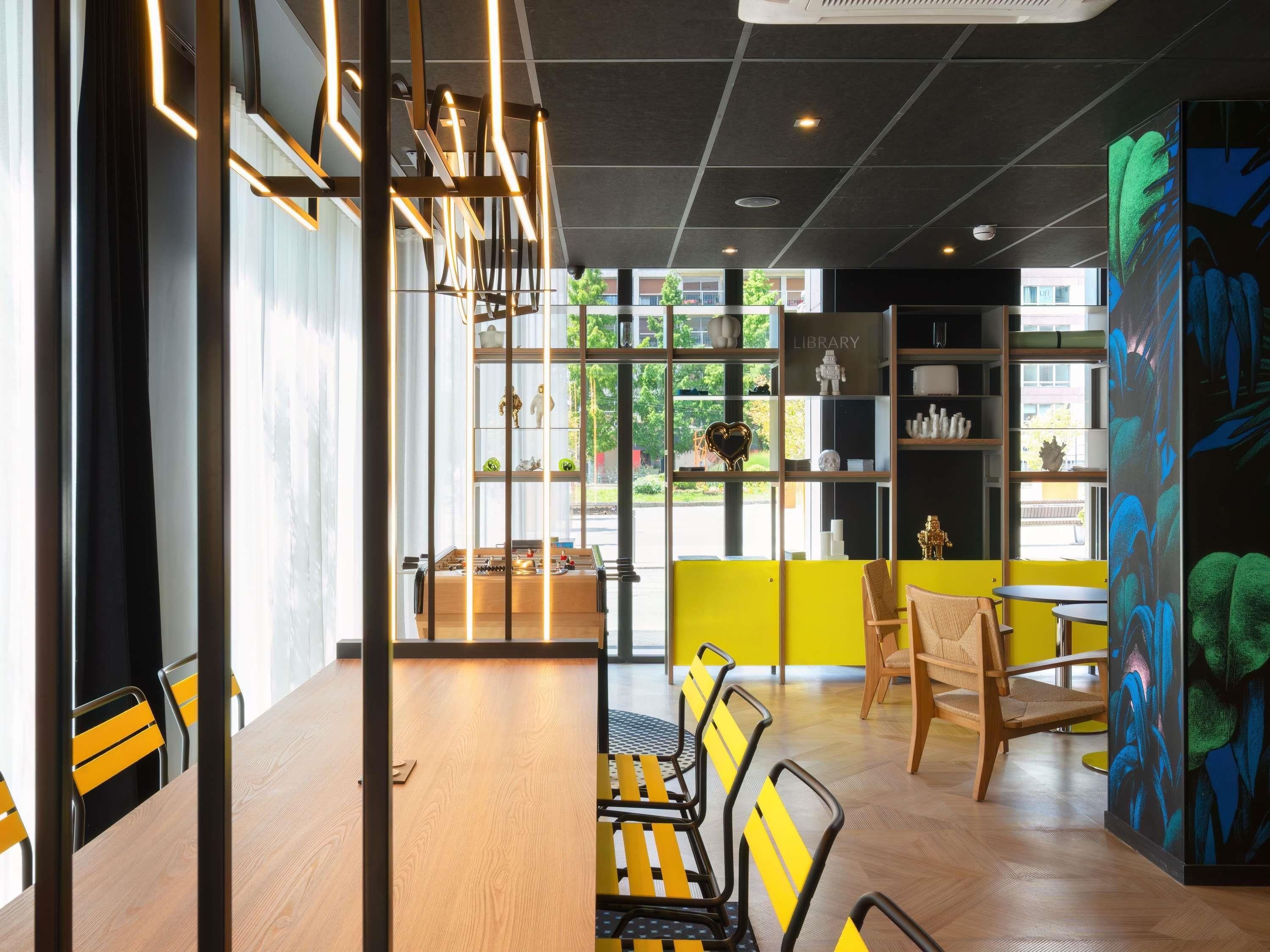 A modern interior with a wooden table and chairs, a bookshelf, and a large window with a view of greenery.