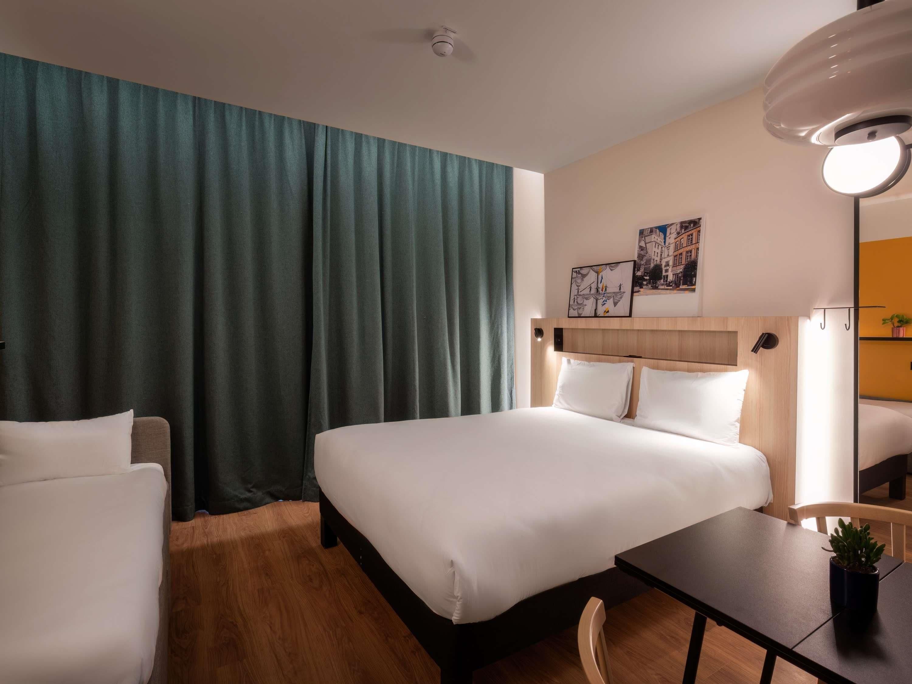 Quarto de hotel com cama, escrivaninha, cadeira, planta em vaso e pintura na parede.