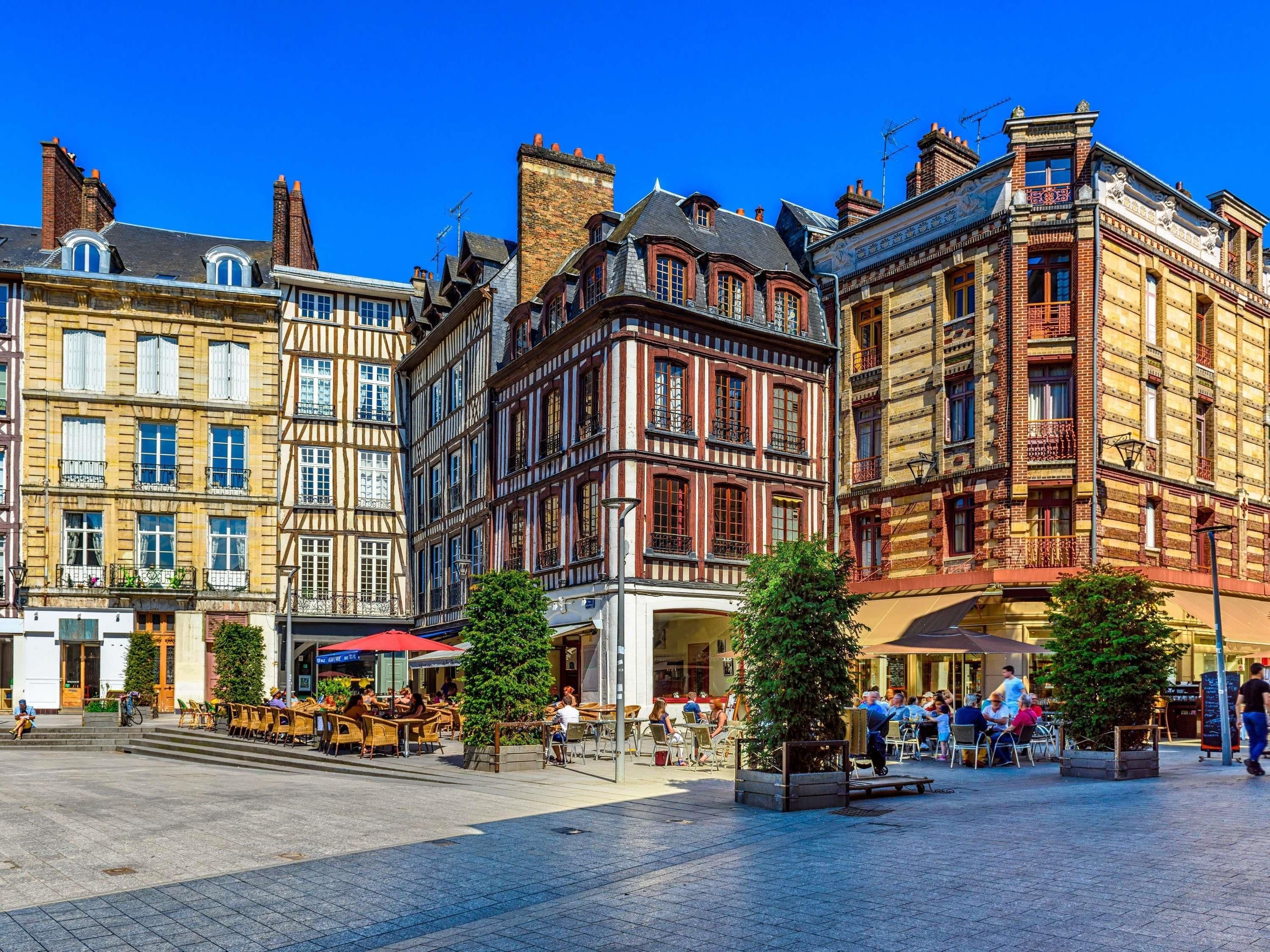 A historic European square with traditional architecture, outdoor seating, and a central tree.