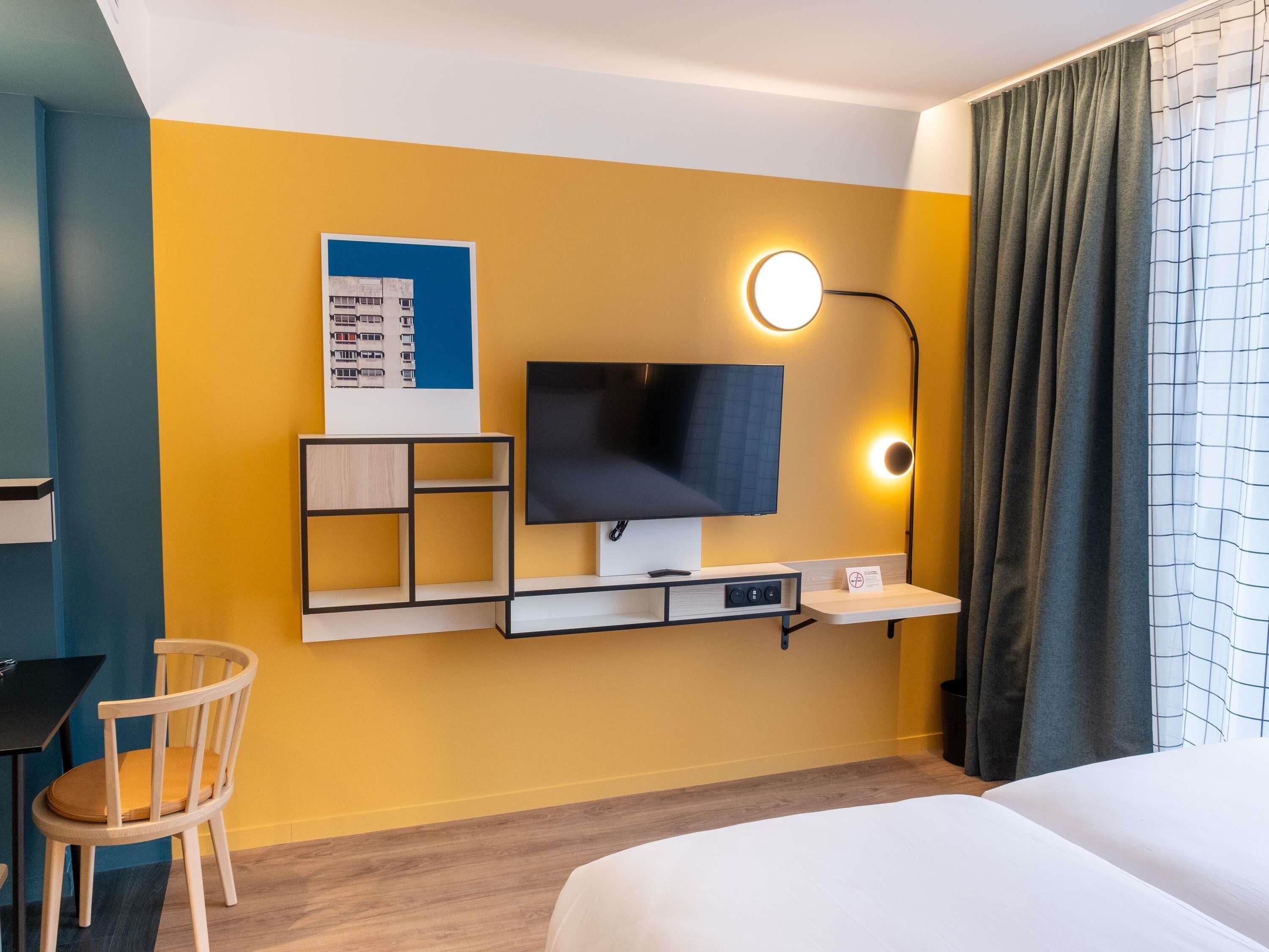 Quarto de hotel com cama, escrivaninha, cadeira, televisão na parede e abajur.