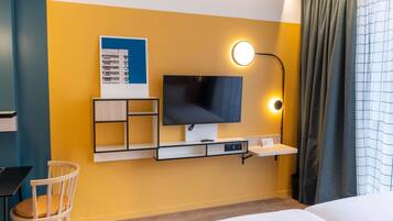 Quarto de hotel com cama, escrivaninha, cadeira, televisão na parede e abajur.