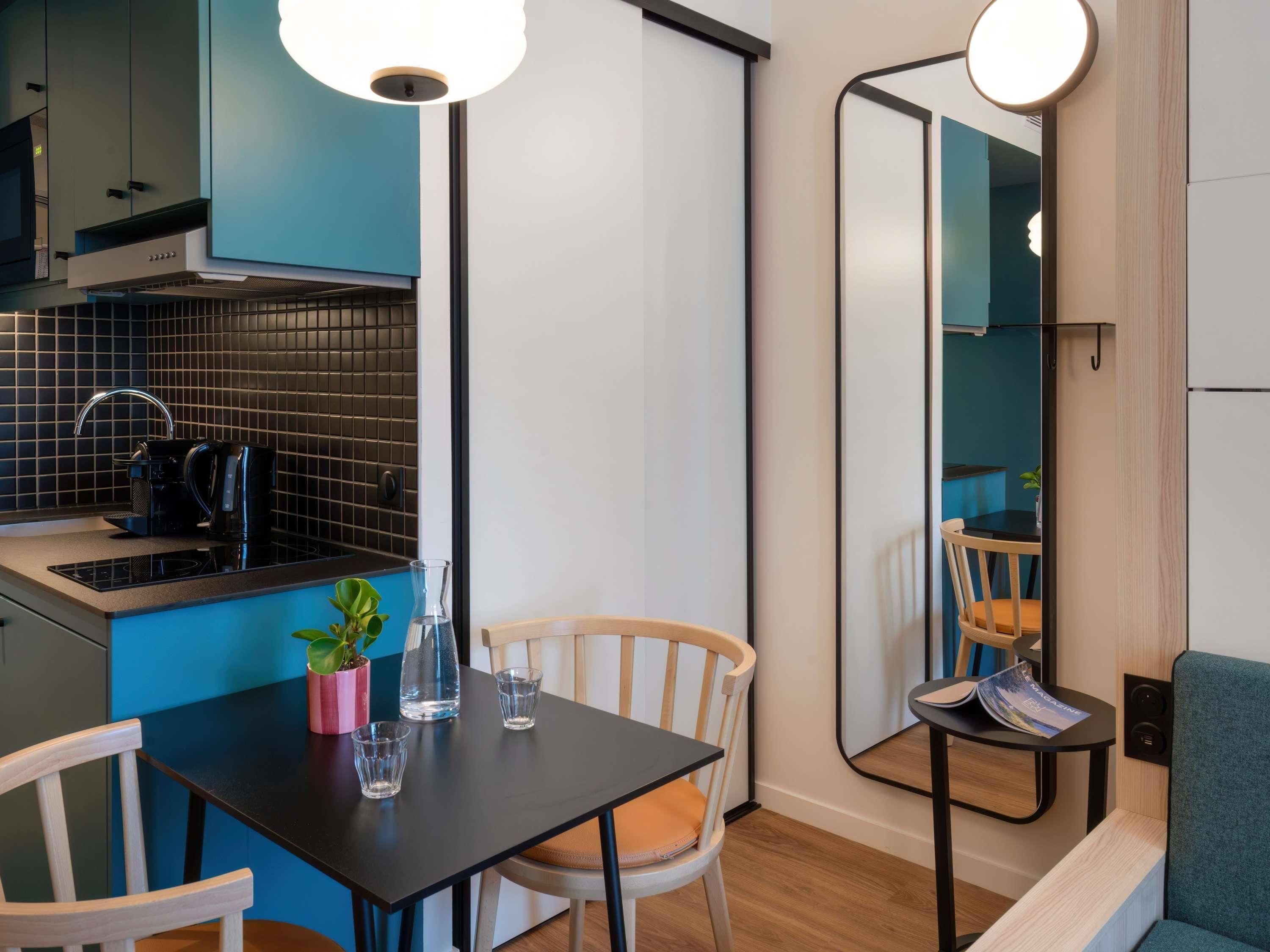 A compact kitchen with a coffee maker, a small table with chairs, a mirror, and a potted plant.
