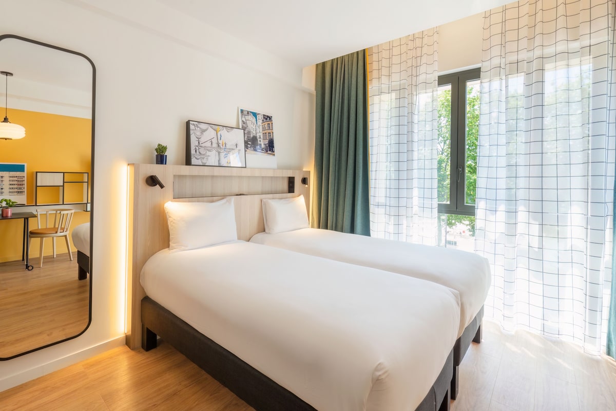 A hotel room with a large bed, a desk with a chair, and a view of greenery through the window.