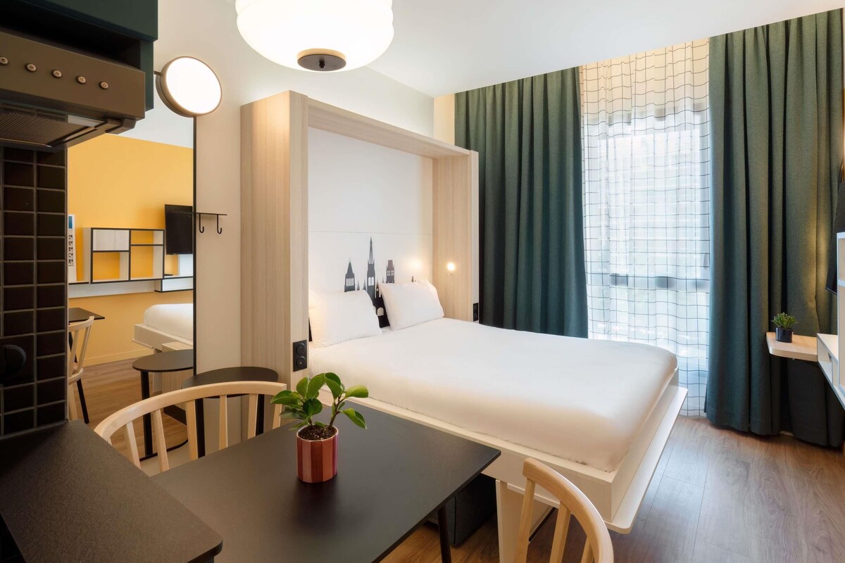 A hotel room with a bed, a desk, a chair, a potted plant, and a large window with curtains.