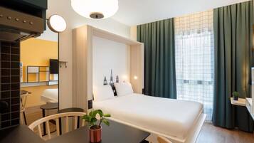A hotel room with a bed, a desk, a chair, a potted plant, and a large window with curtains.