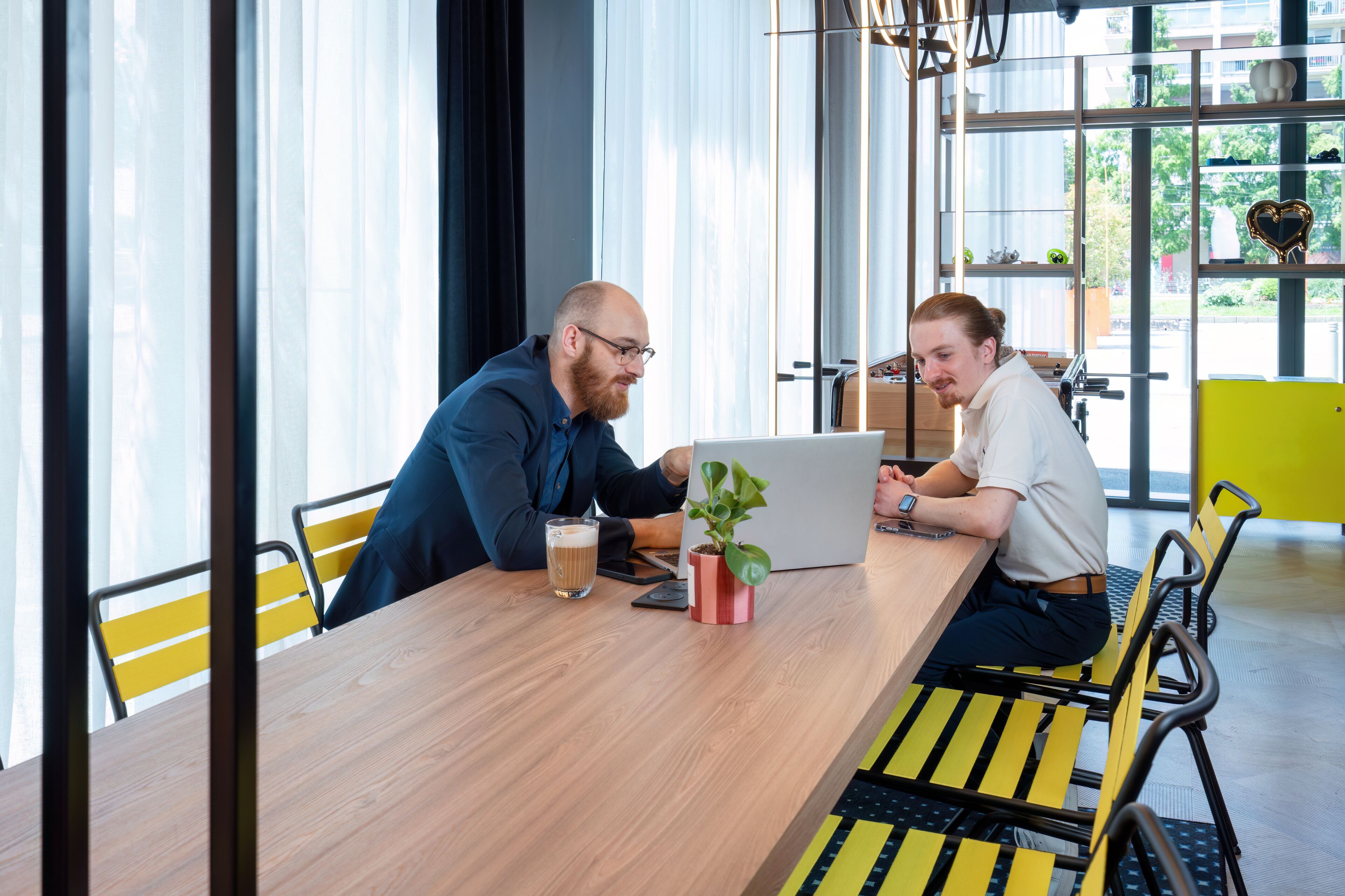 Twee personen aan een houten tafel met een laptop, een biertje en een potplant.