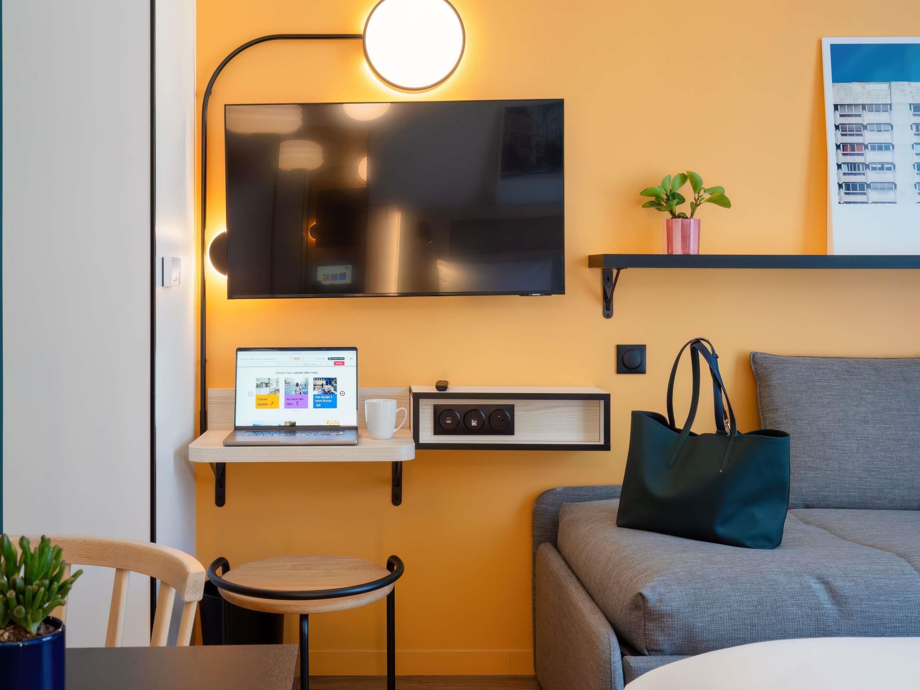 Une chambre moderne avec un téléviseur à écran plat, un ordinateur portable, une tasse à café, une radio, une plante et un sac de couleur turquoise sur un canapé gris.