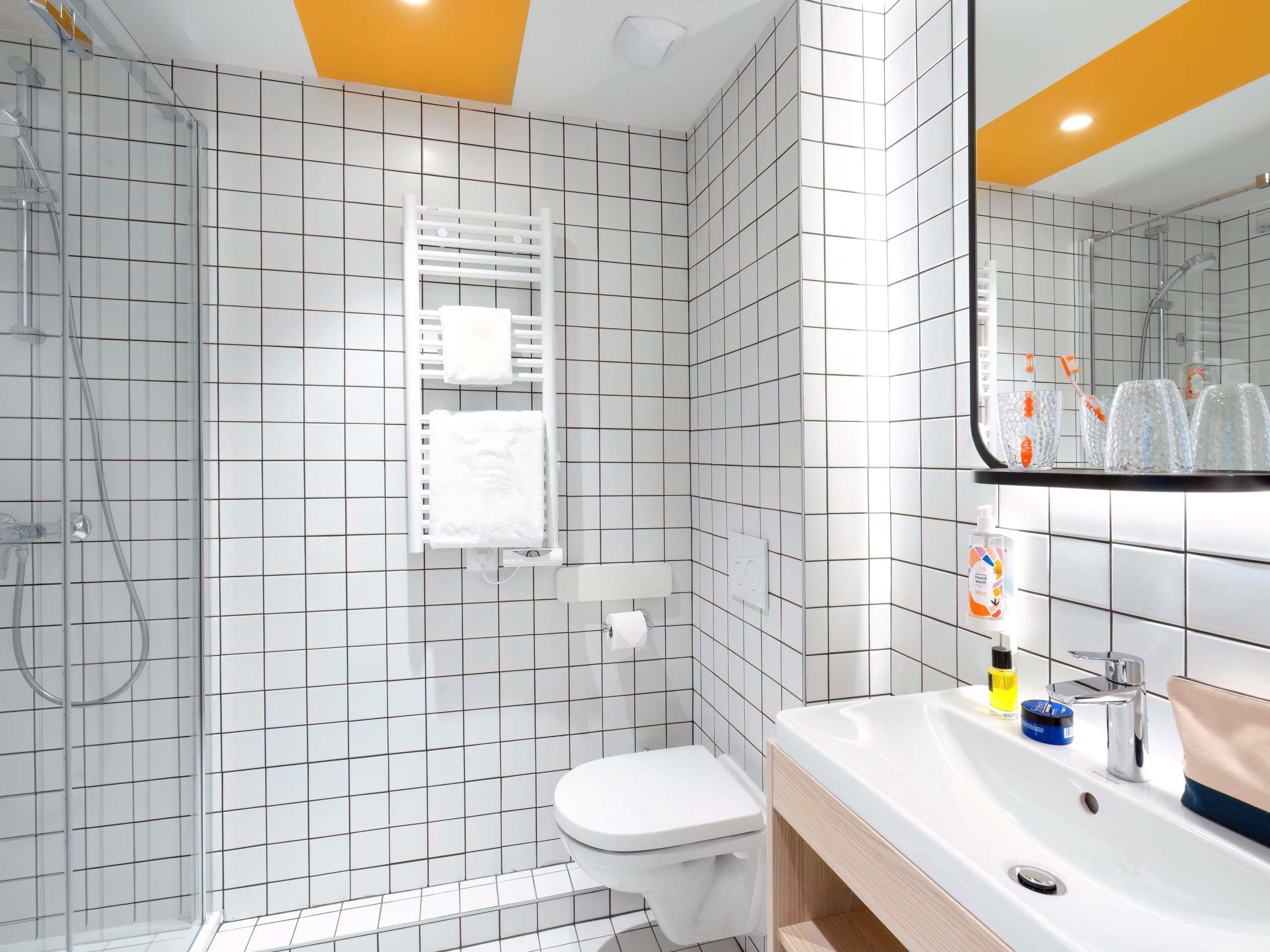 A modern bathroom with white tiled walls, a glass shower enclosure, a white toilet, and a sink with a mirror above it.