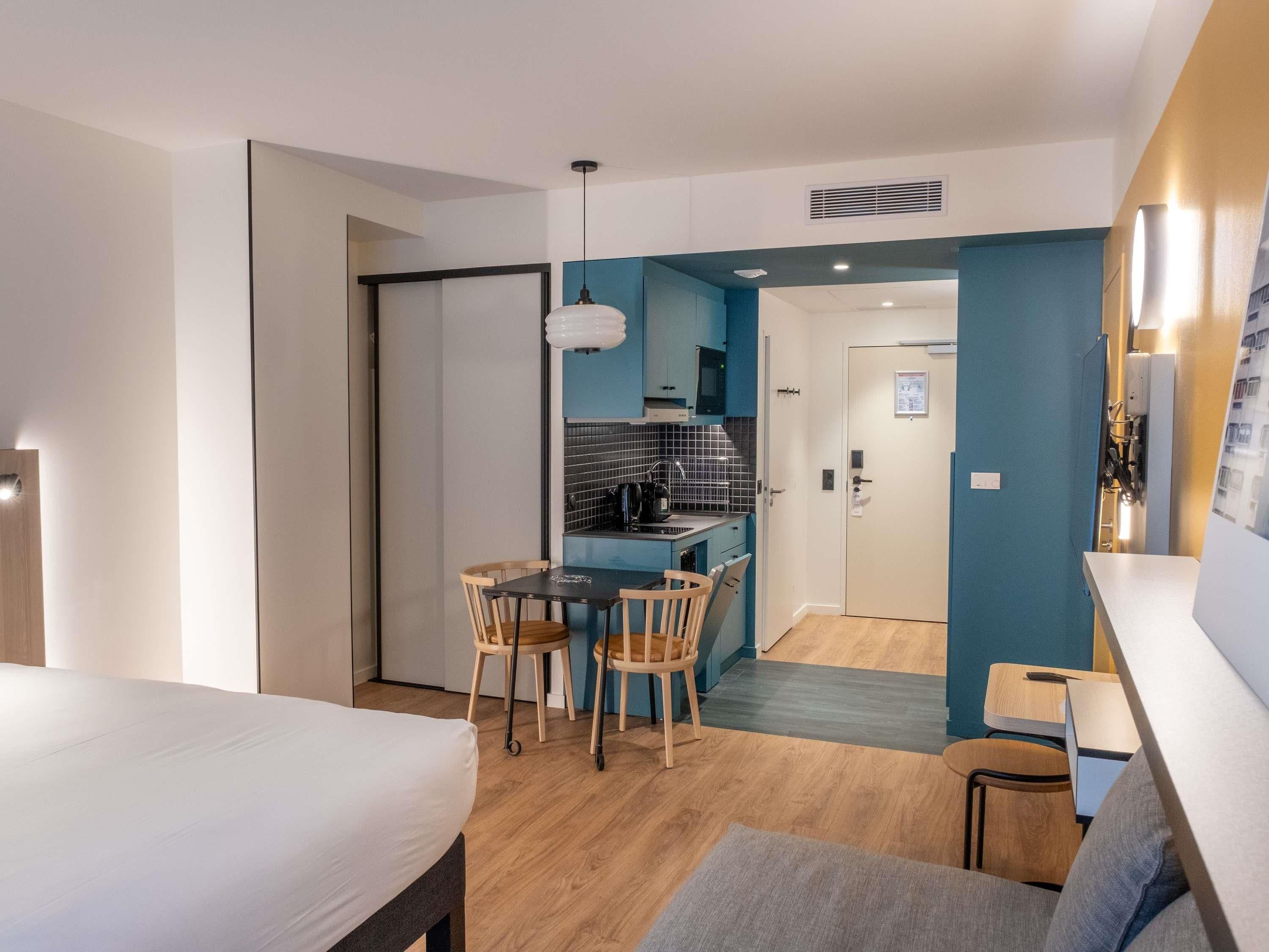 A modern hotel room with a bed, a dining area with chairs, a small kitchen, and a bathroom.
