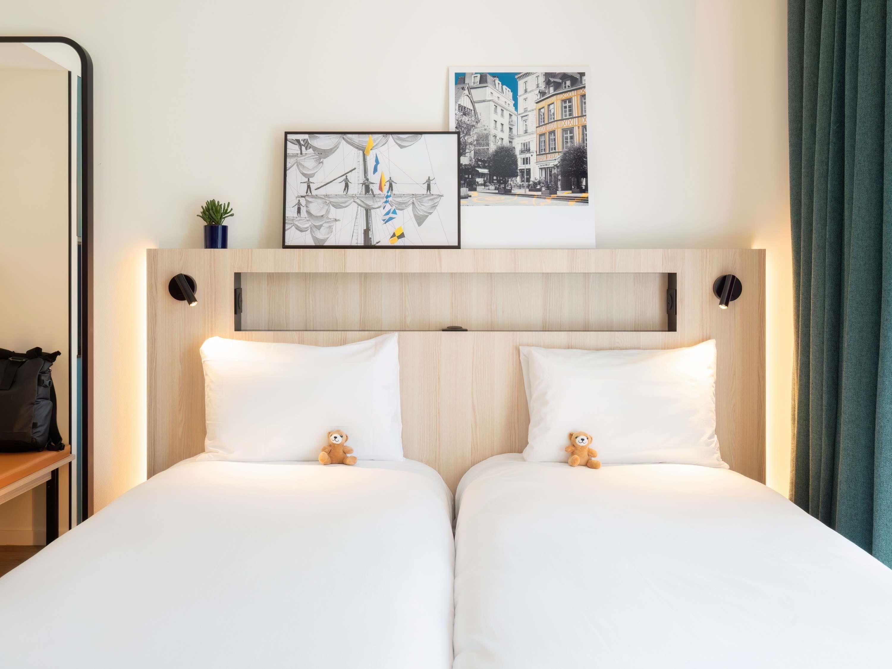 Une chambre d’hôtel avec une tête de lit en bois, deux lits avec du linge de lit blanc, deux ours en peluche, une petite plante et deux tableaux encadrés.