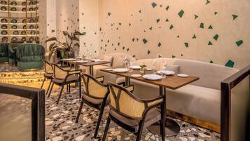A modern dining area with a white sofa, wooden tables, and chairs. There are decorative wall tiles and a patterned floor.