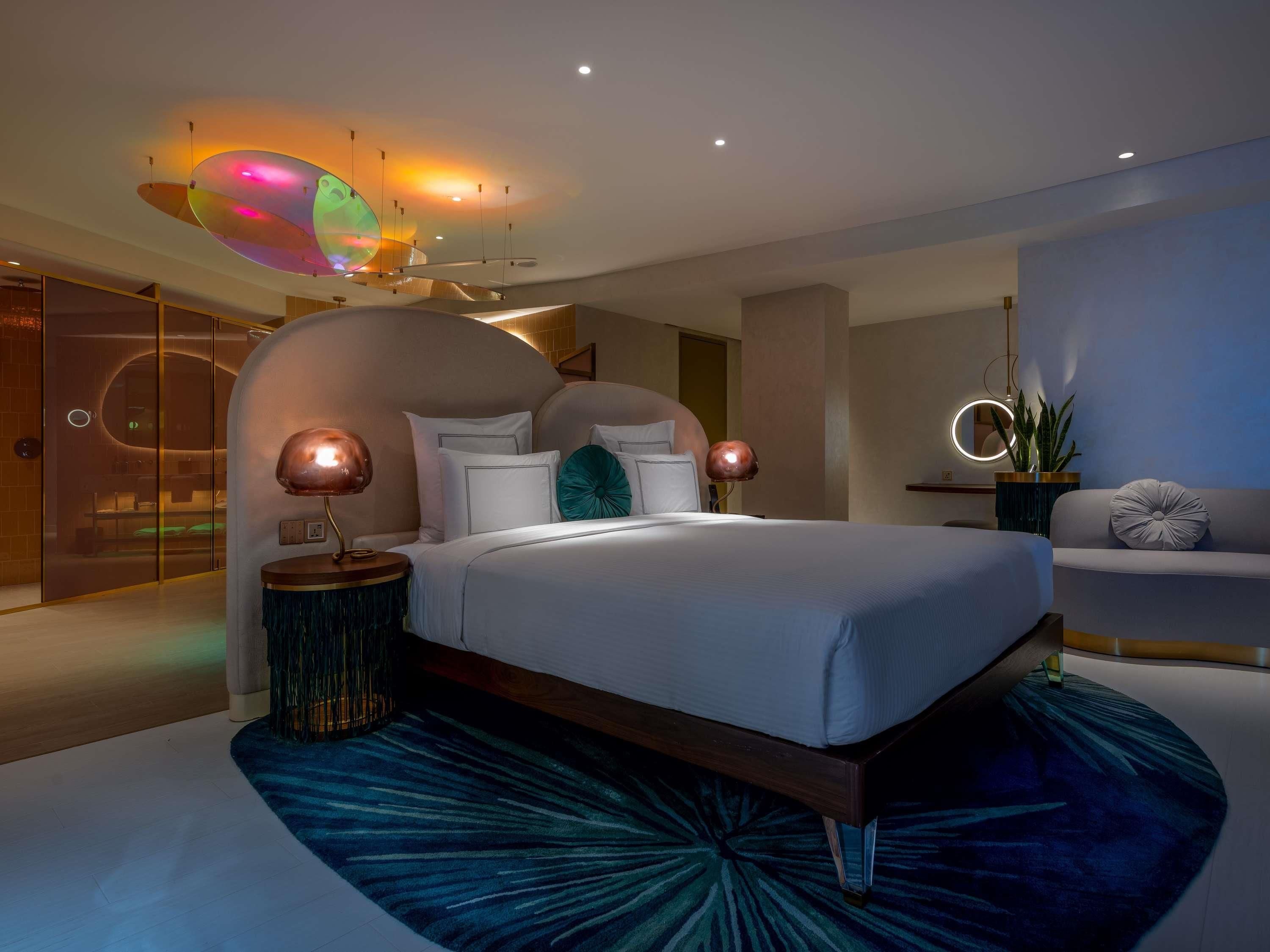 A modern bedroom with a curved headboard, a large bed, a side table, a sofa, and a unique ceiling light fixture.