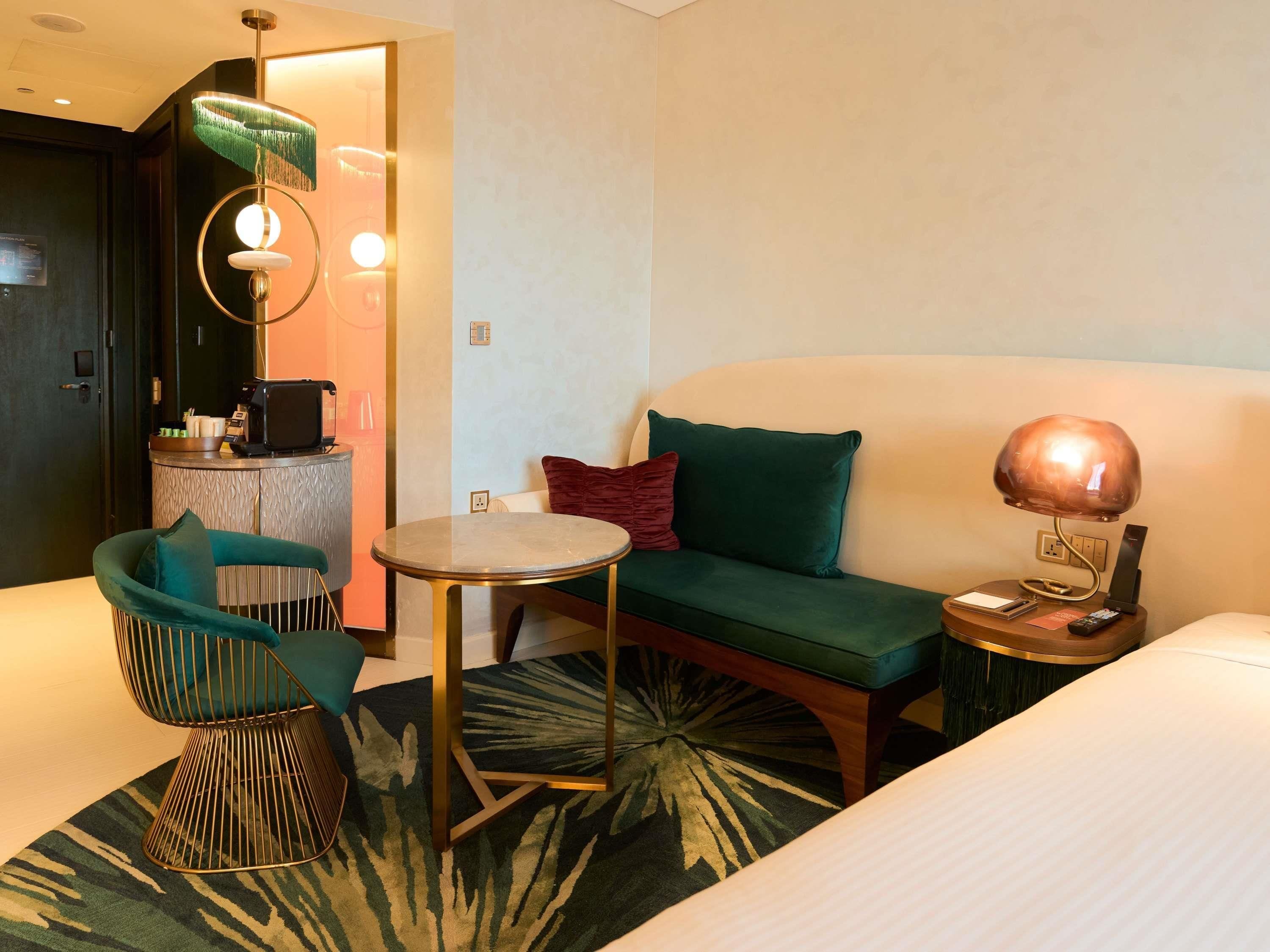 Habitación de hotel con cama, una mesa redonda, sofá, sillón, lámpara, escritorio con teléfono y control remoto, y una alfombra pequeña.