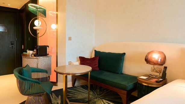 A hotel room with a bed, a round table, a sofa, a chair, a lamp, a desk with a phone and a remote, and a small rug. - Gem Forest Hotel Nairobi - MGallery Collection (Nairobi)