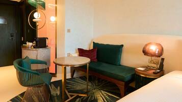 Habitación de hotel con cama, una mesa redonda, sofá, sillón, lámpara, escritorio con teléfono y control remoto, y una alfombra pequeña.