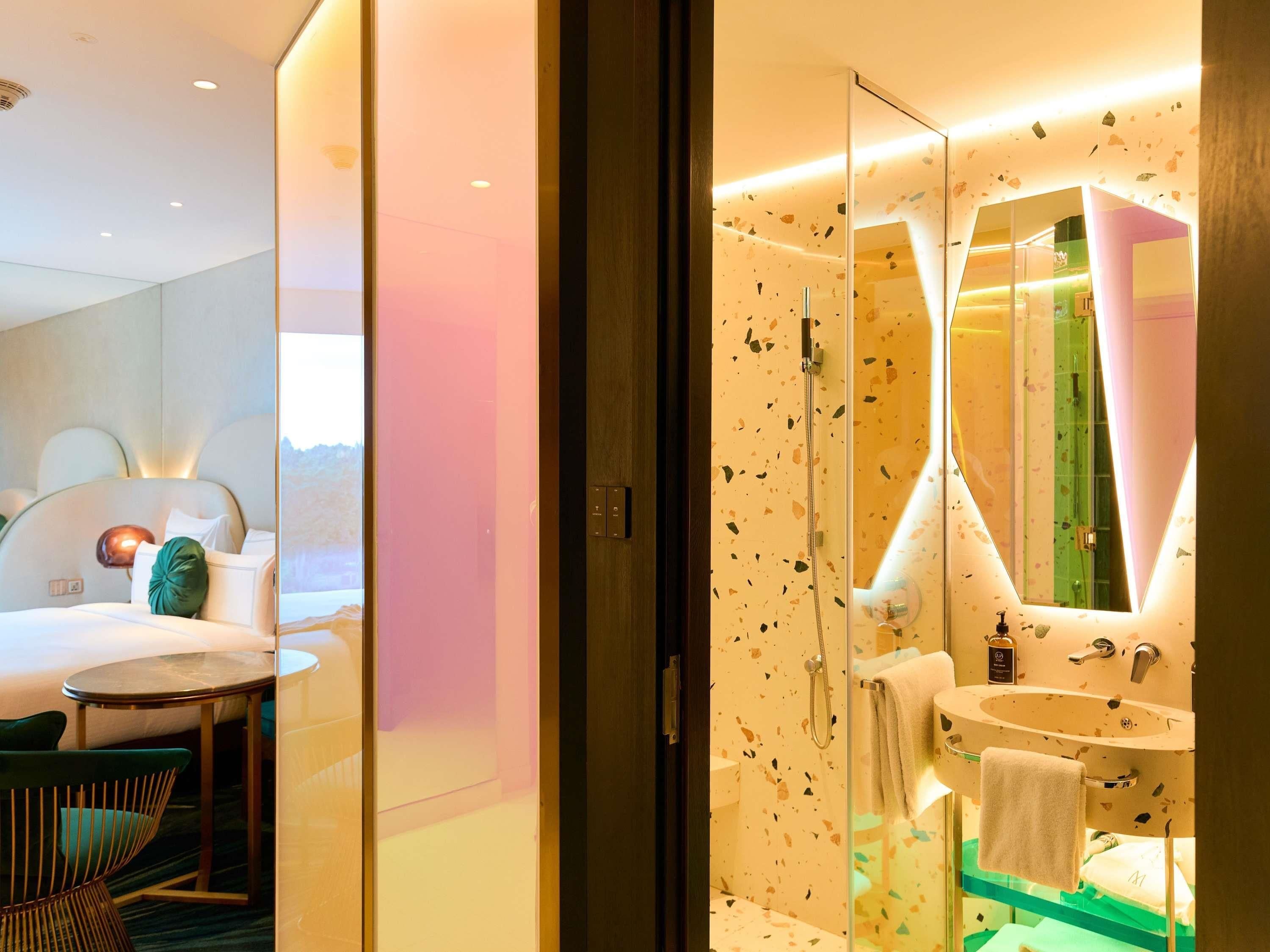 Una habitación de hotel moderna con cama, una mesa redonda, zona de ducha con mampara de cristal y baño con lavamanos y espejo.
