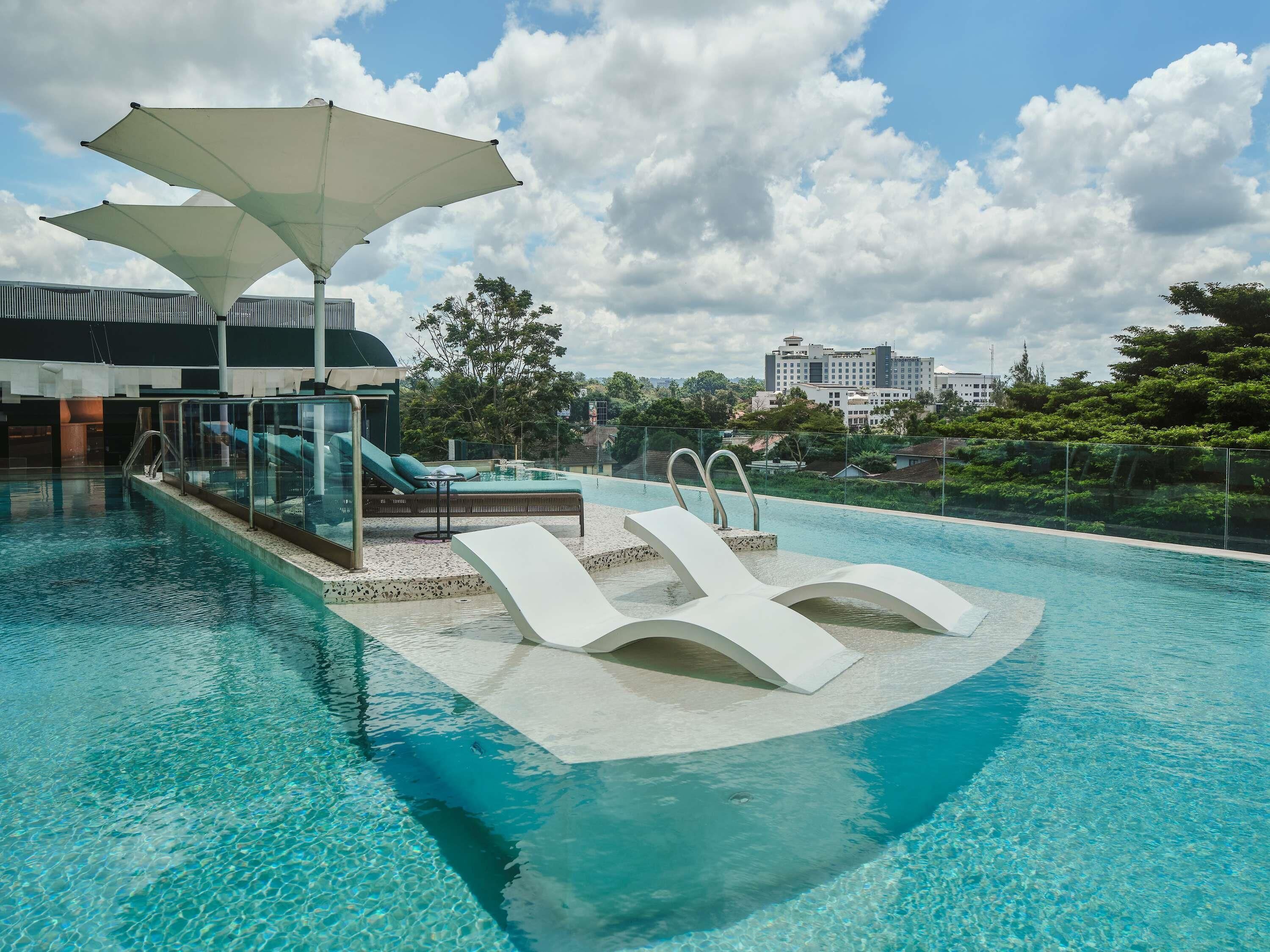 A swimming pool with lounge chairs and umbrellas, a glass balustrade, and a view of a cityscape in the background.