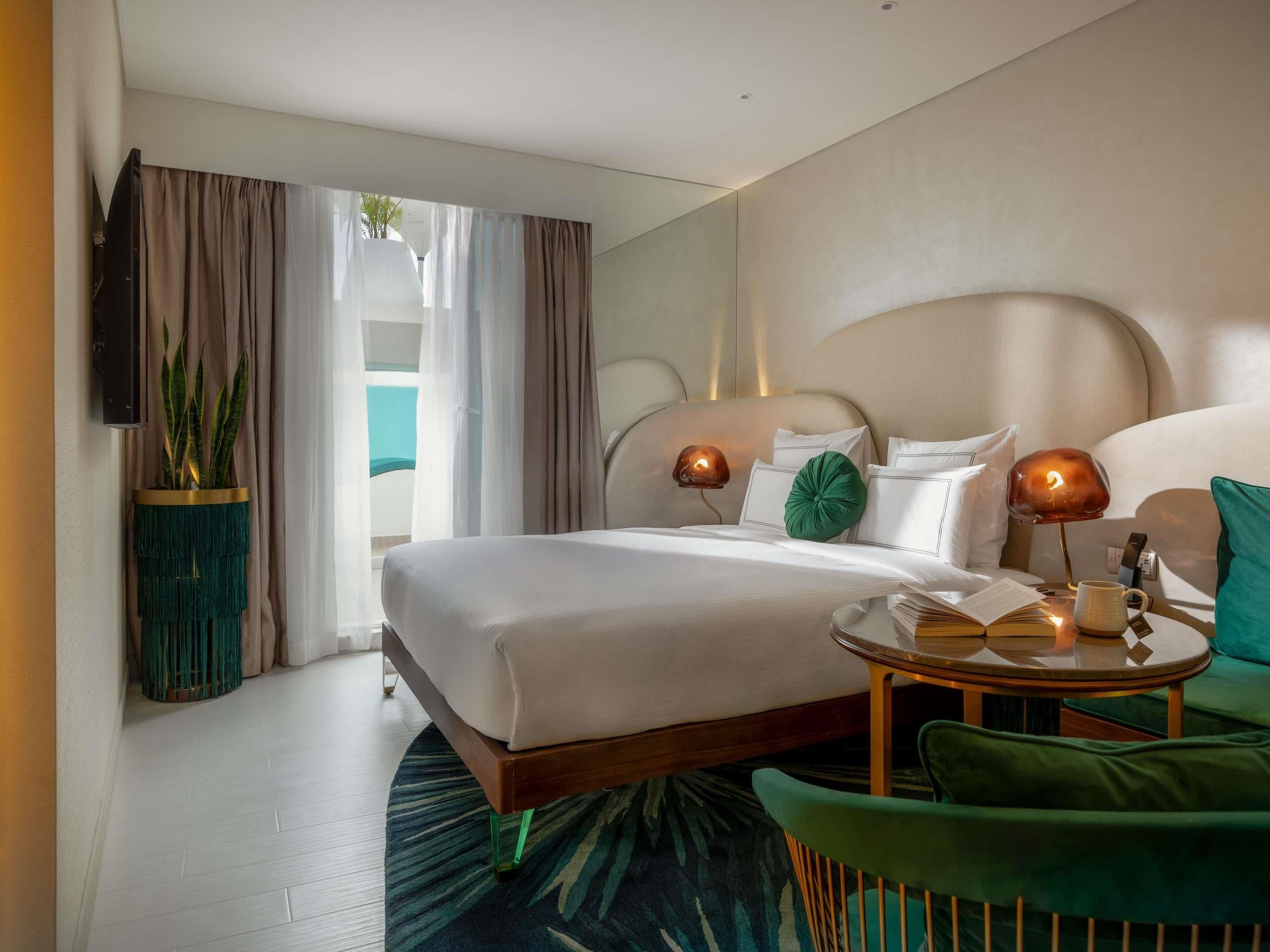 A hotel room with a bed, bedside lamps, a small table with a book and a mug, a green chair, and a plant in a decorative pot.
