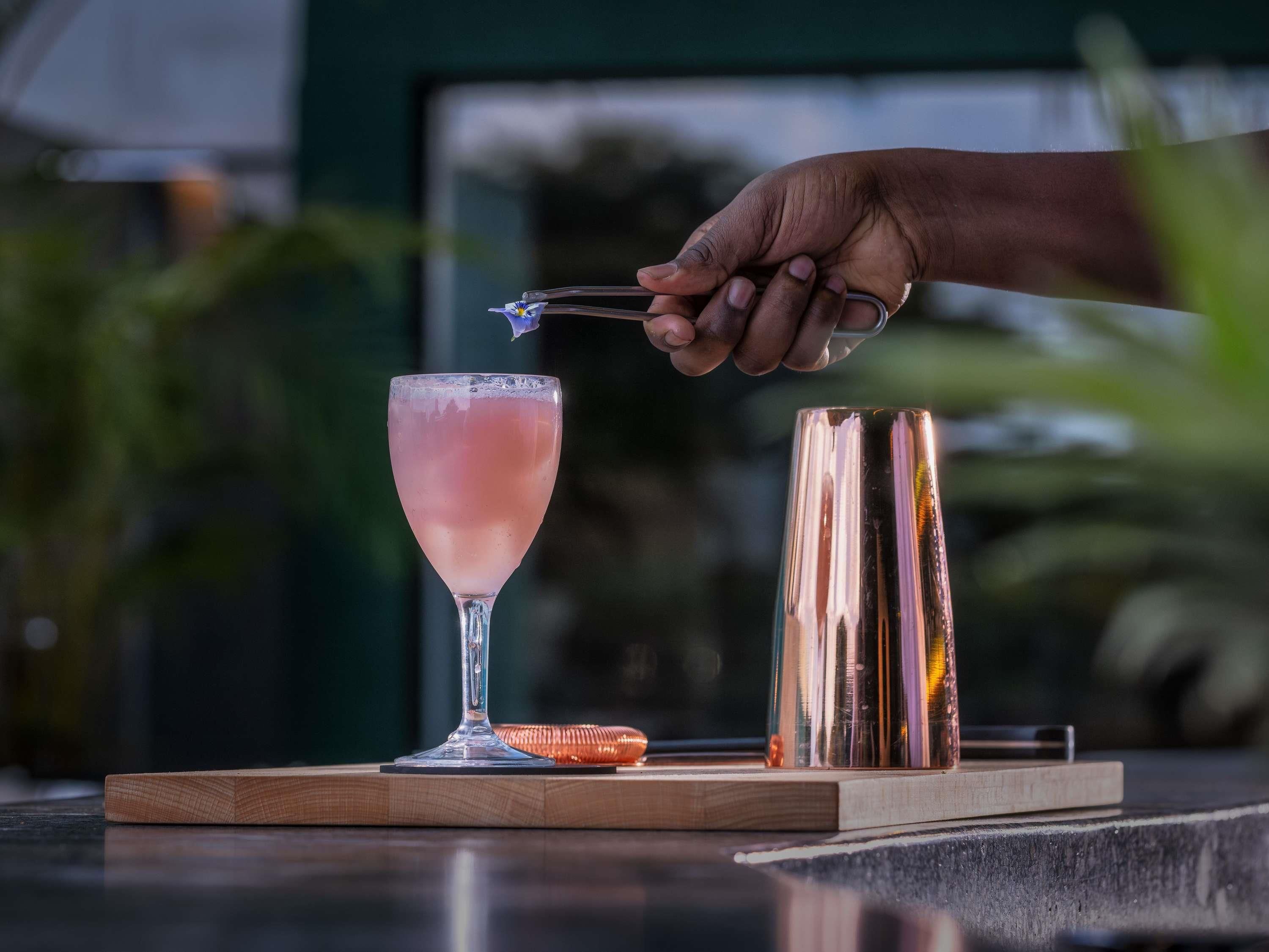 A hand holding a cocktail shaker, a glass with a pink drink, and a copper shaker on a wooden board.