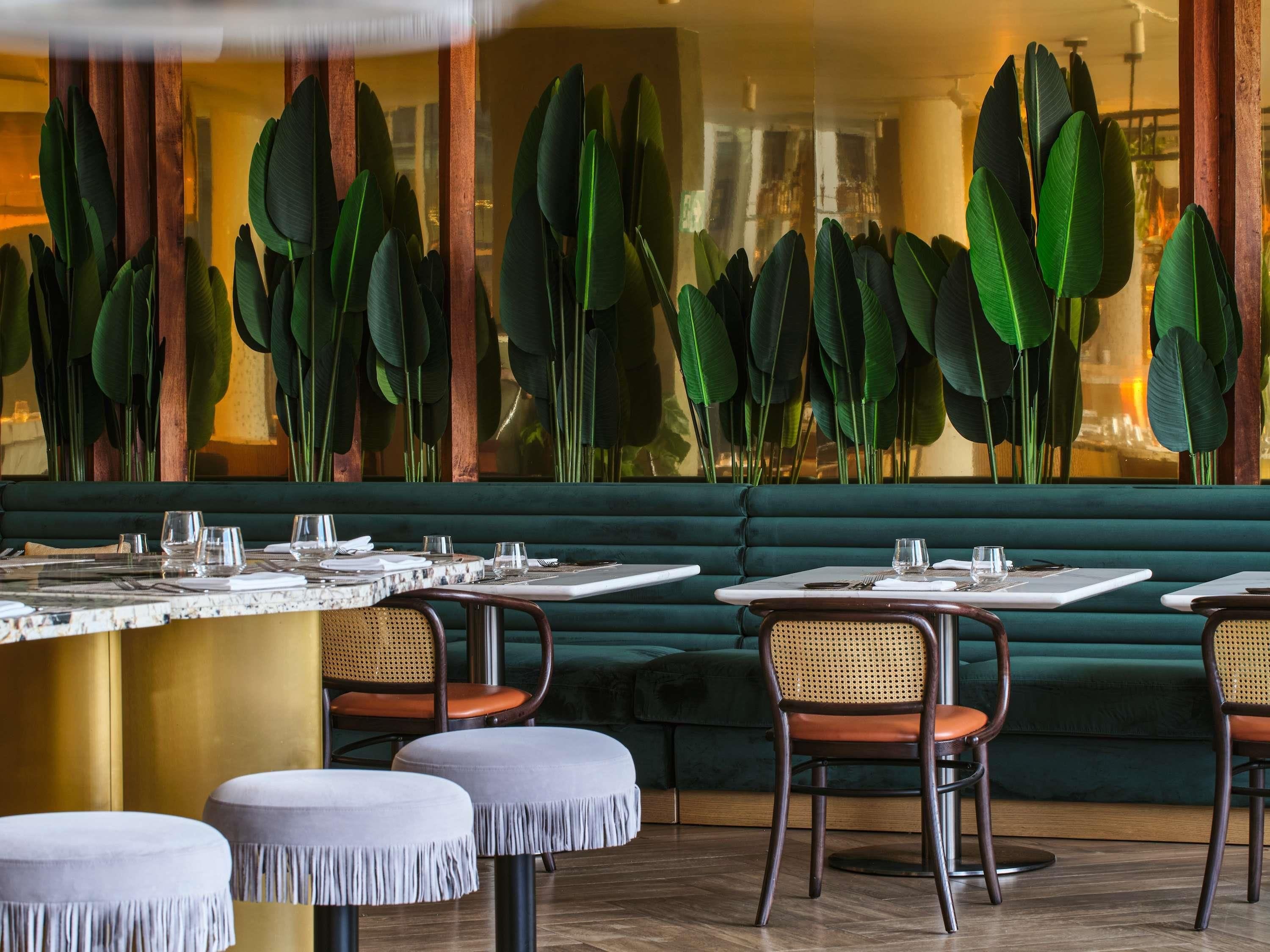 A restaurant interior with a green booth seating area, round tables with white placemats, and large green leaves as a decorative wall feature.