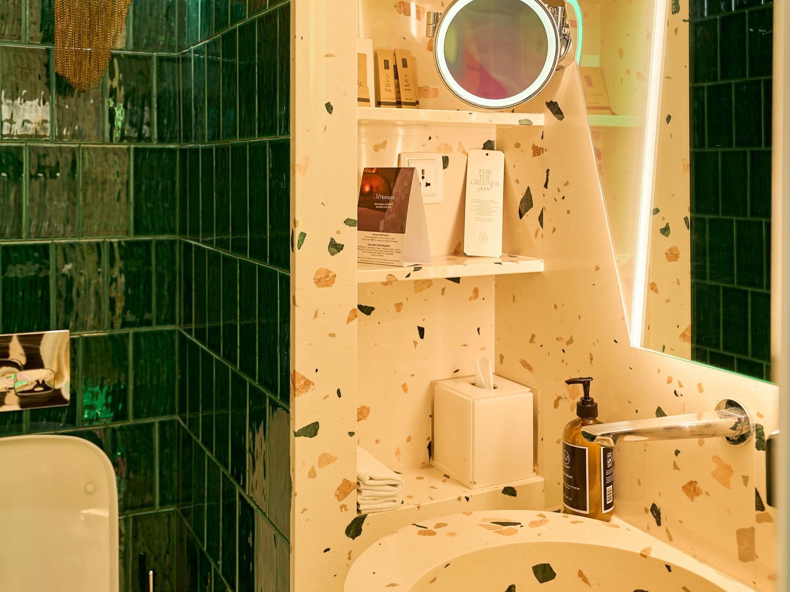 A bathroom with a green-tiled wall, a round mirror, shelves with toiletries, and a sink.