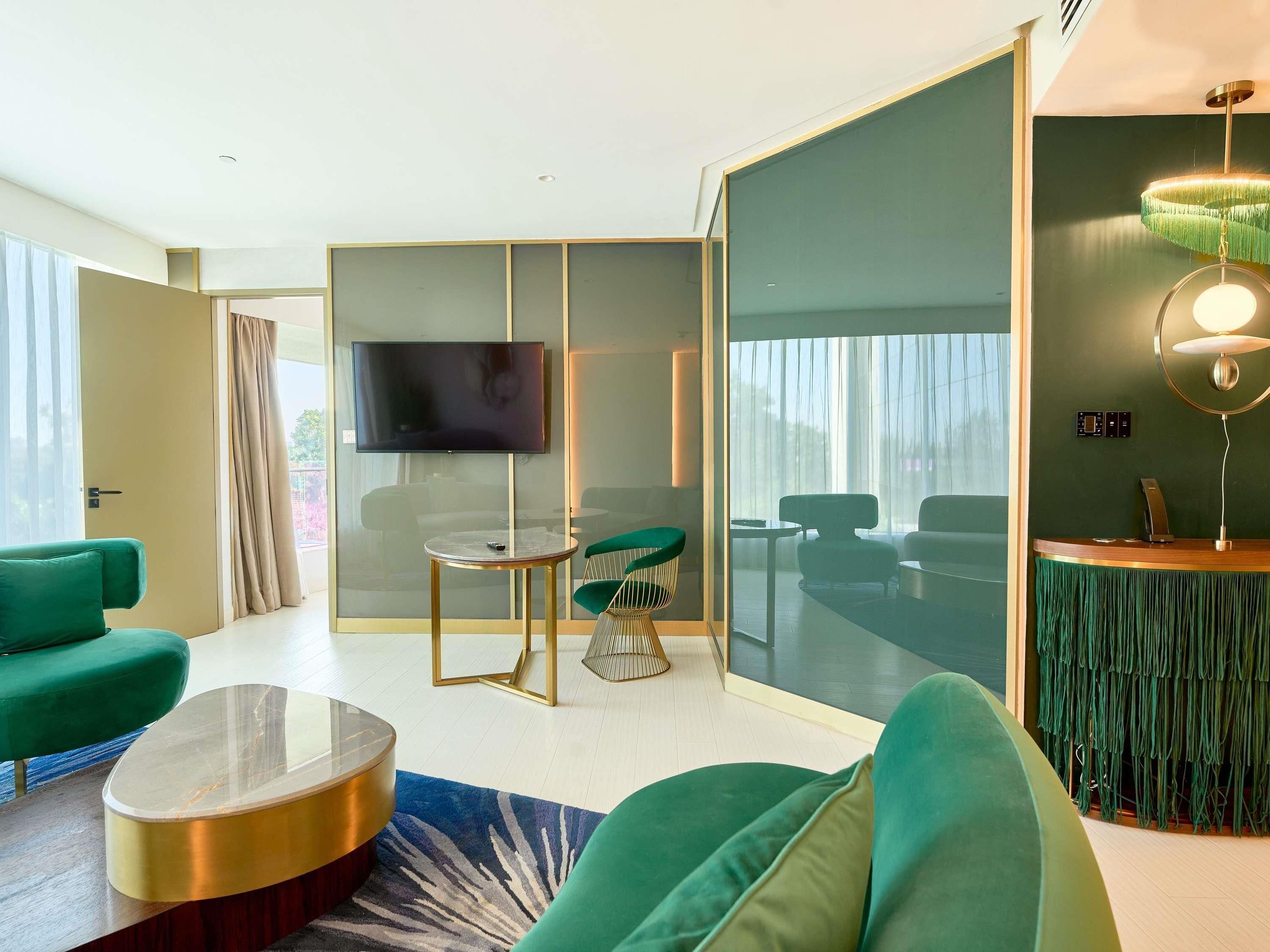 A modern living room with a flat-screen TV, green sofas, a round coffee table, and a large window with curtains.