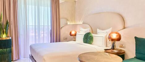A hotel room with a large bed, bedside lamps, a round table, a green sofa, and a potted plant.