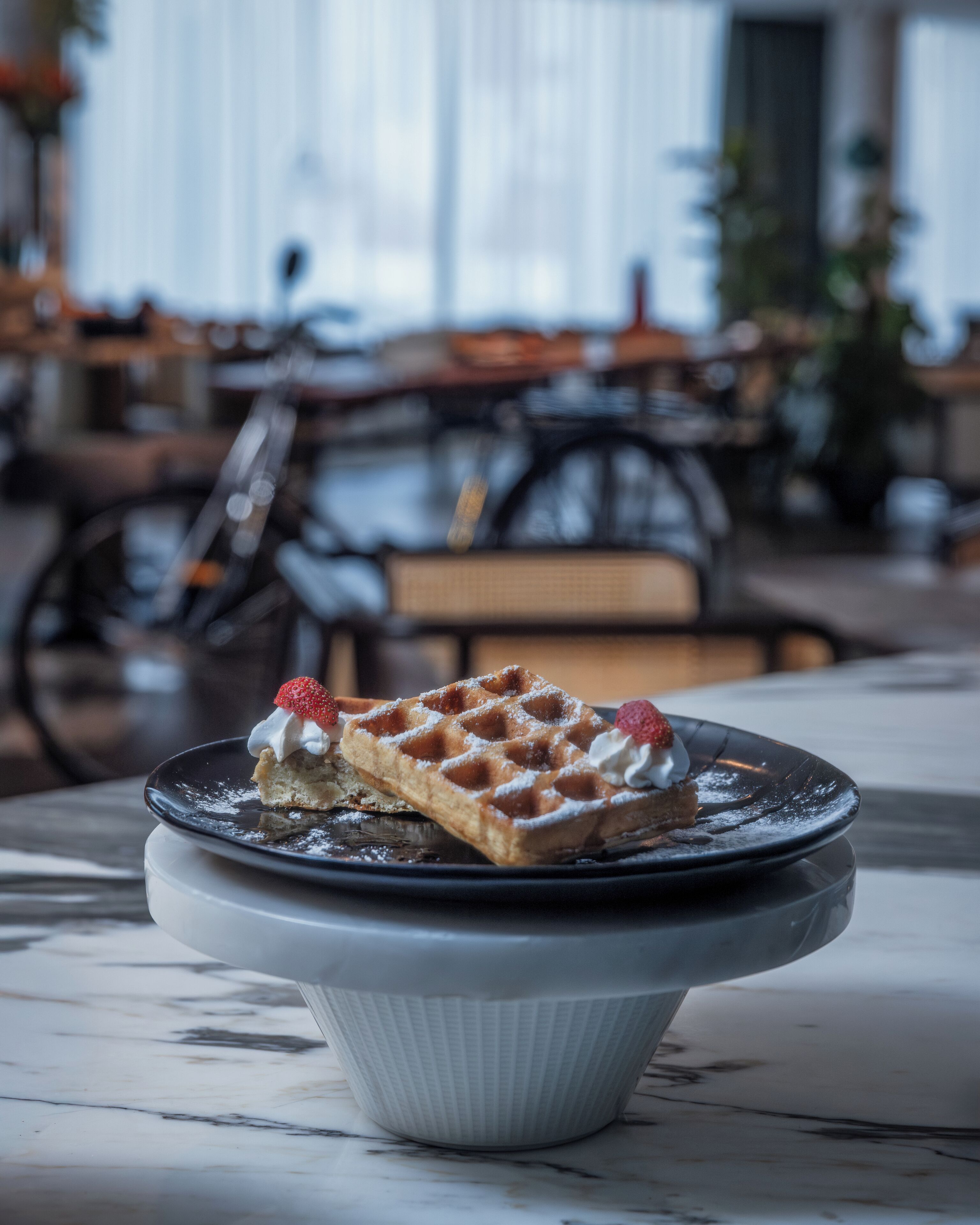 A waffle with whipped cream and a strawberry on a plate, placed on a marble table in a restaurant setting.