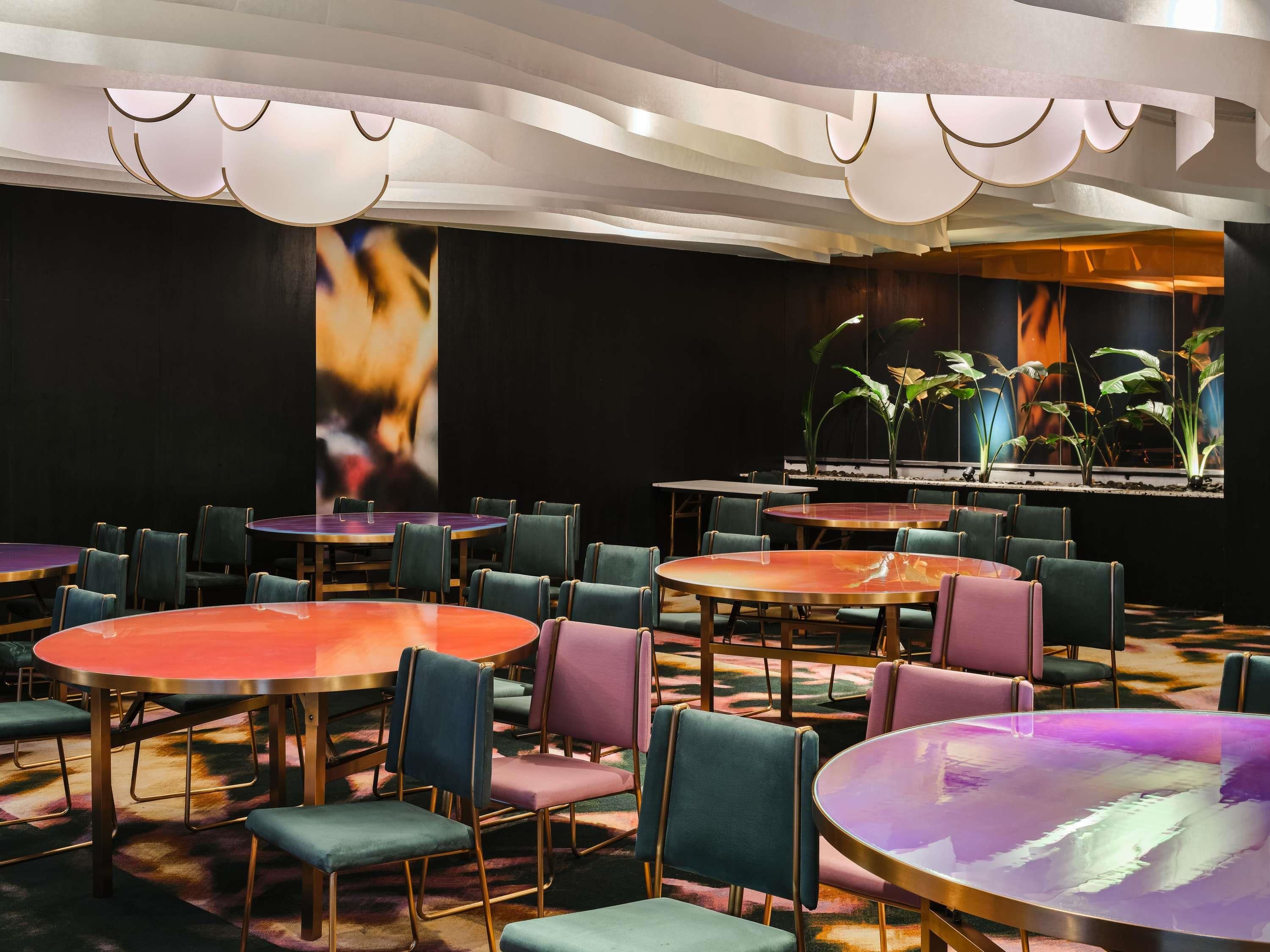 A hotel room with multiple round tables and chairs, a large screen displaying a fire, and a plant wall.