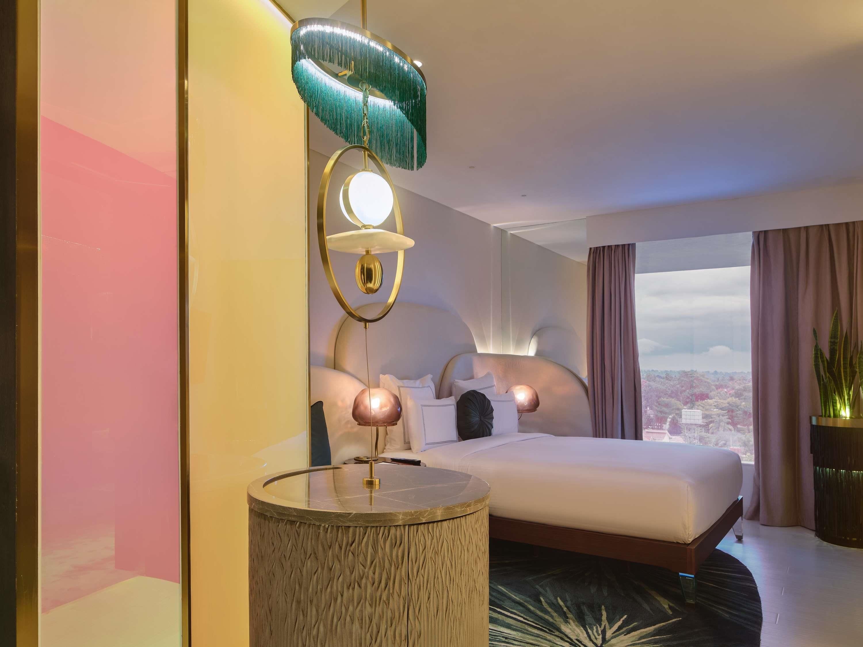 A modern hotel room with a bed, bedside lamps, a round table, and a view of the city.