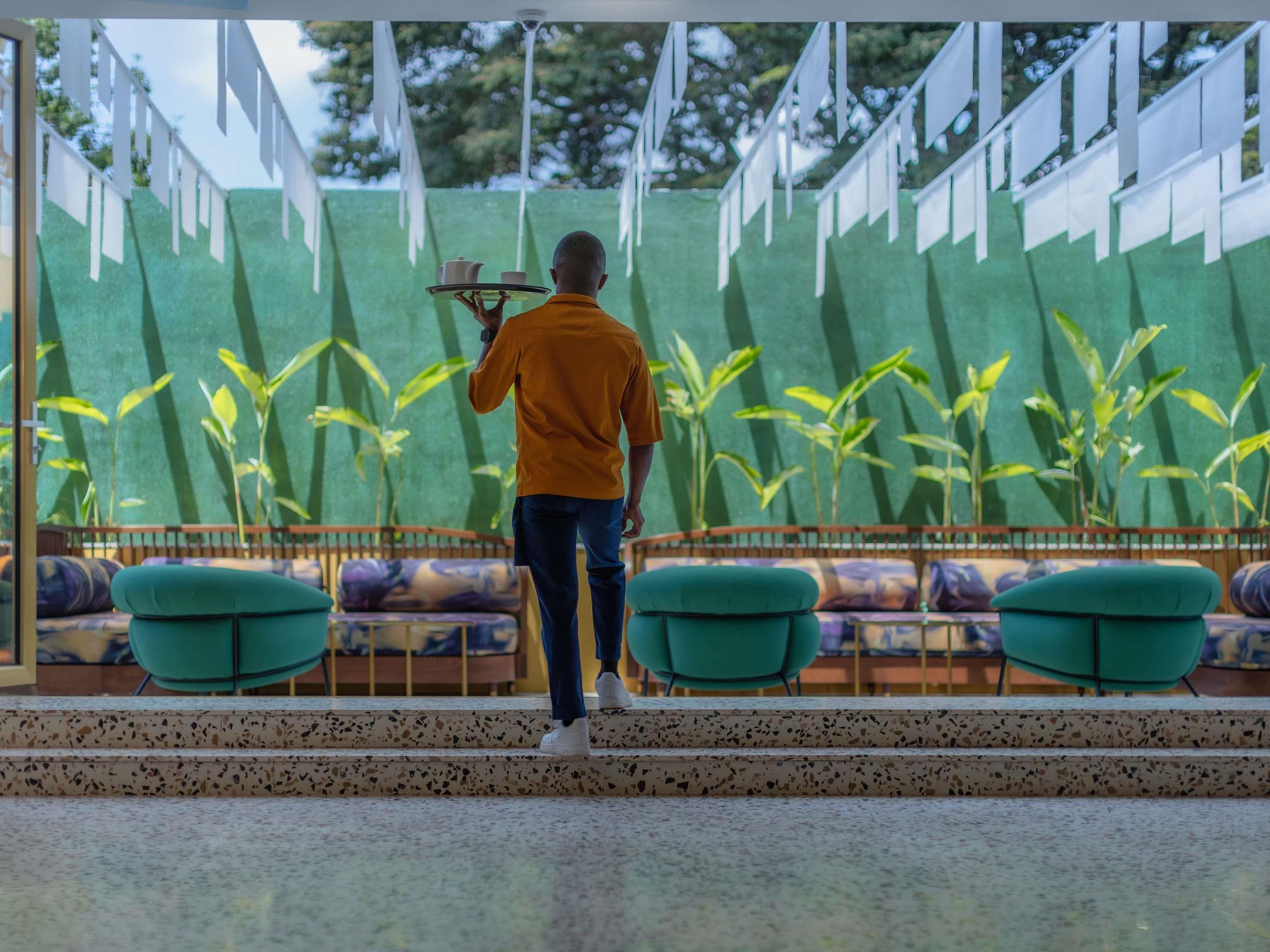 A person standing on a balcony with a tray of dishes, a green wall with vertical stripes, and a seating area with green chairs.