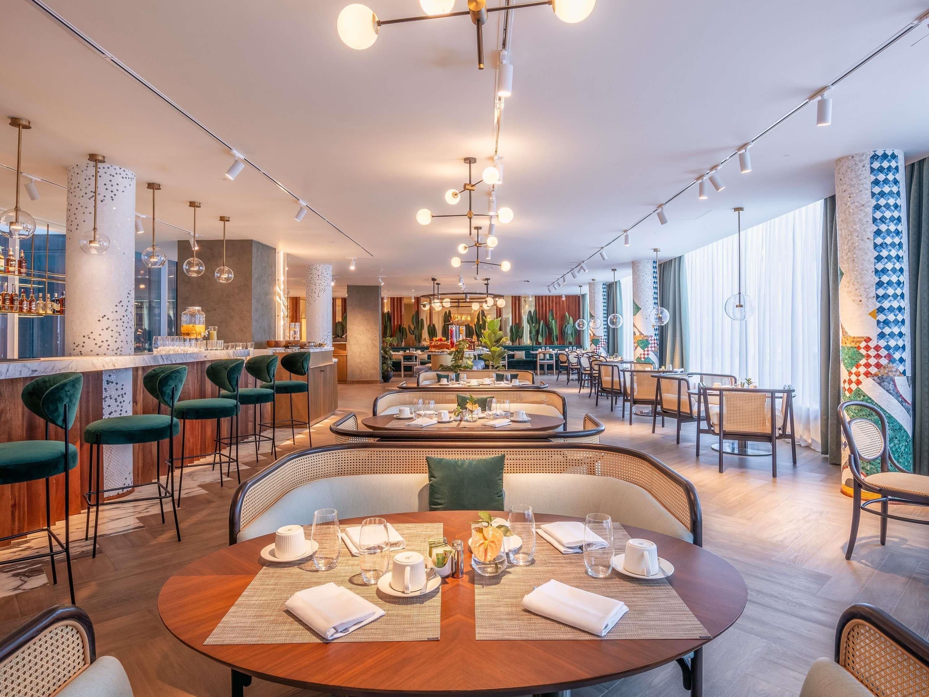 A modern hotel dining area with round tables, bar seating, and a central bar area.