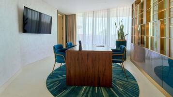 Una sala de reuniones moderna con una mesa de madera, sillas azules, un televisor grande y un ventanal con cortinas translúcidas.